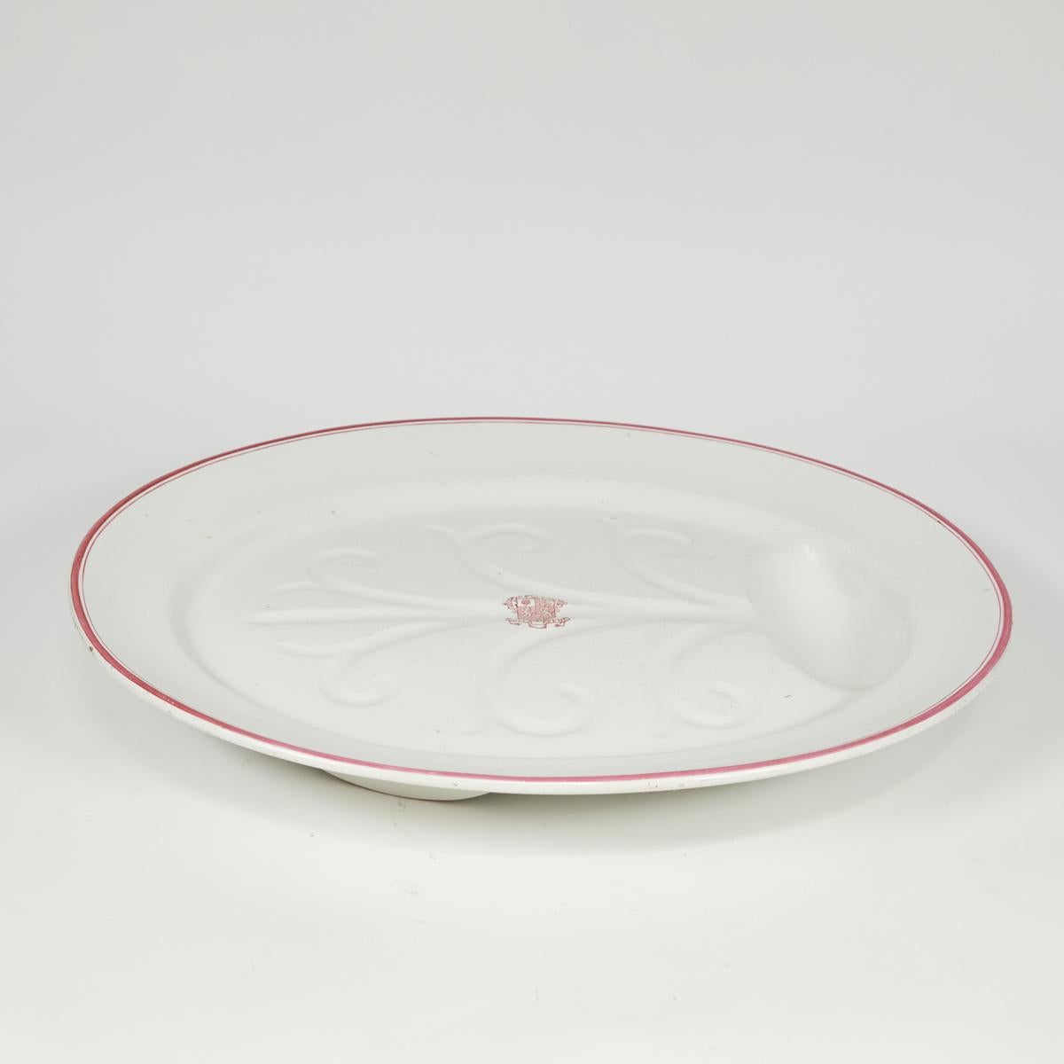 English late 19th-century white porcelain oval serving platter with delicate pink trim. The base features a small indentation to catch sauce and drippings, and is accented by a simplified palmette carving. An elegant rose-colored armorial insignia