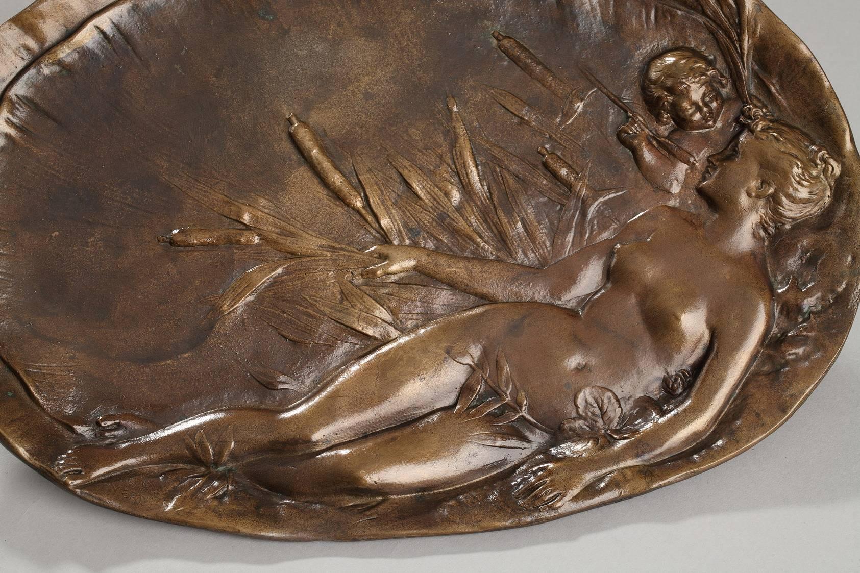 Oval platter in patinated bronze decorated with a sculpted, bas-relief woman stretched out among reeds and cattails. She is nude, and behind her, a young cupid is gazing at her face her through the reeds. This scene, saturated with the sensuality of