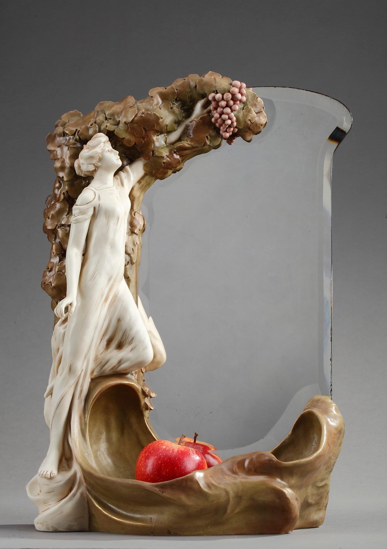 This original Art Nouveau mirror was crafted of polychrome porcelain. The beleved, rectangular glass is decorated with a young woman picking a grape. The lower part of the mirror forms a receptacle. Marked underneath: Royal Dux Bohemia and numbered