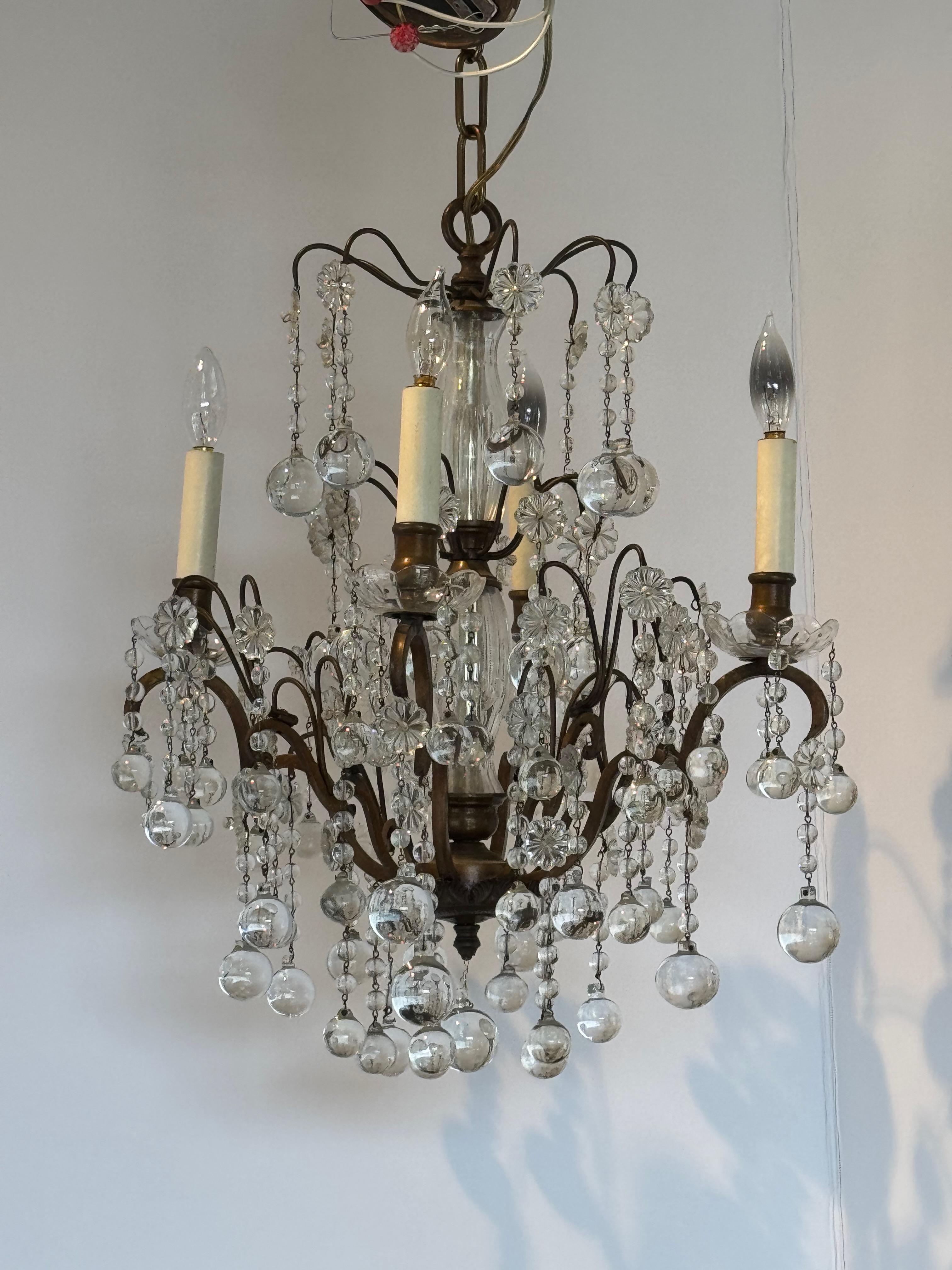 Lampadario in bronzo ornato con gocce di cristallo a cascata e rosette floreali. I bracci graziosamente arcuati sostengono luci a candela, completate da una colonna centrale in cristallo. Con un raffinato equilibrio di metallo e vetro, questo pezzo