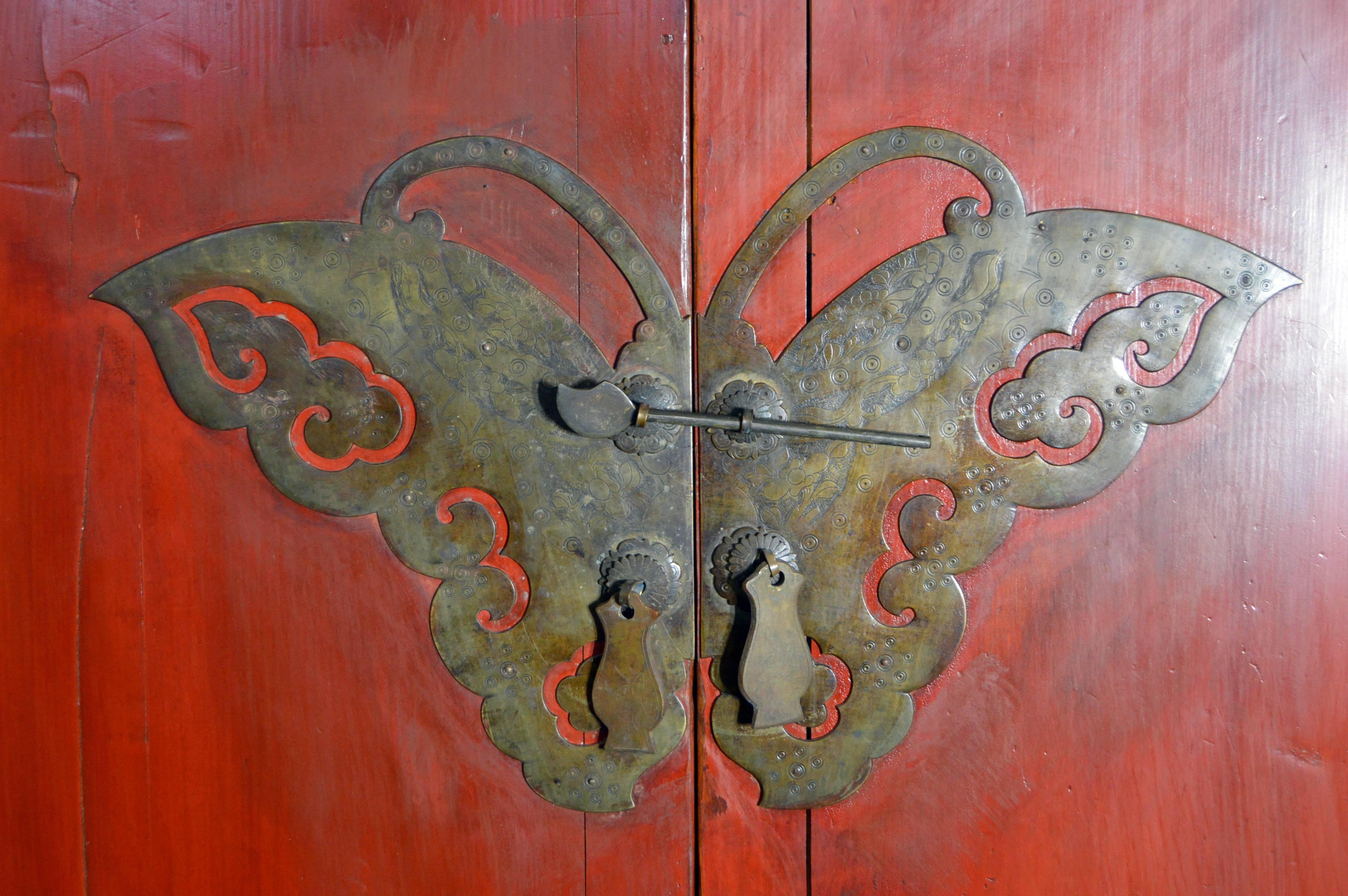 A Chinese two-door cabinet from the late 19th century, with a red lacquer finish, hidden drawers, carved skirt and brass butterfly hardware. This Chinese cabinet features a linear silhouette, raised on four straight legs, while below the doors sits