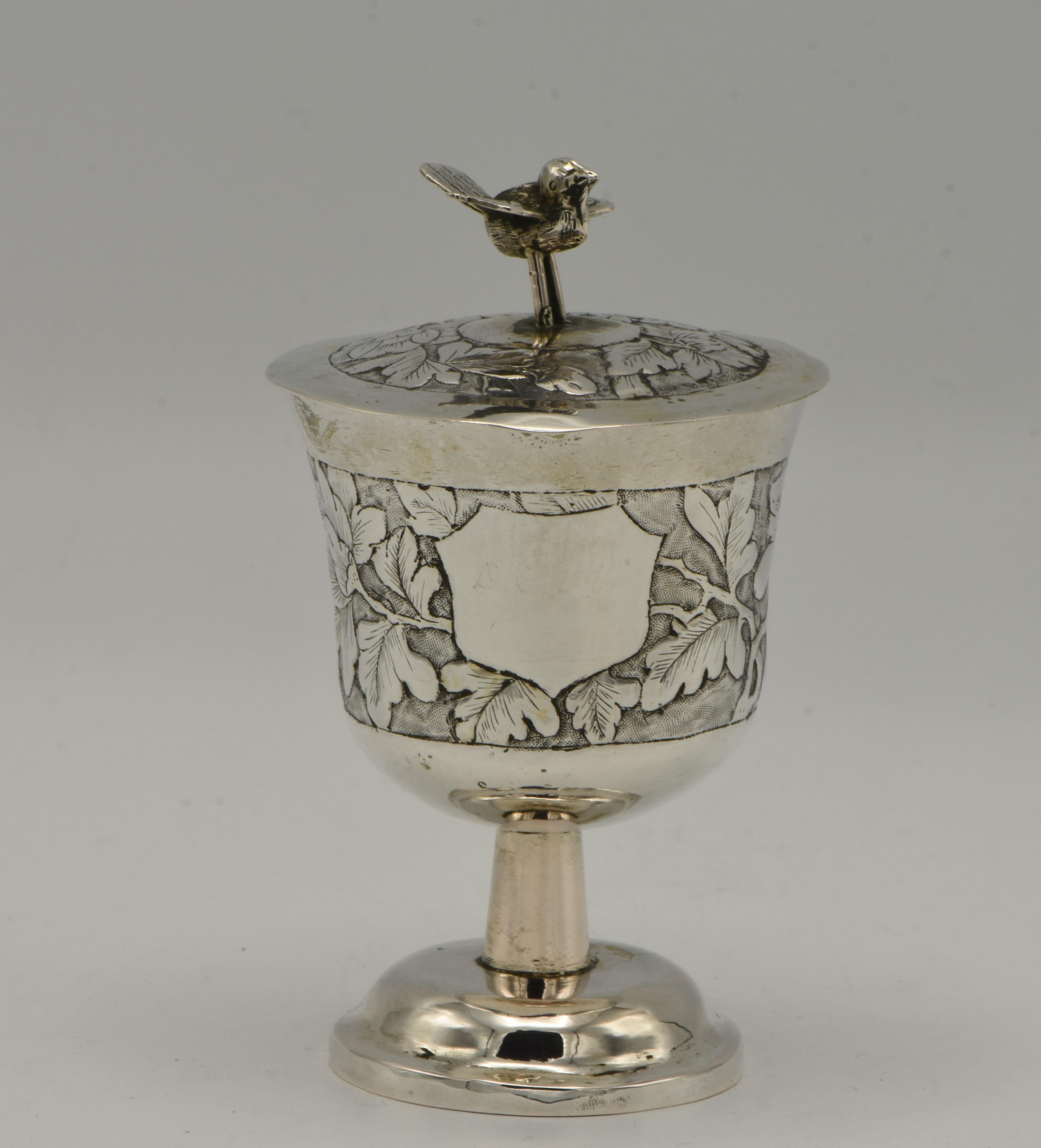 Chinese export silver covered Kiddush goblet, 19th century. 
Round cup on a stem mounted to a flat circular foot. Stepped domed cover with bird finial. With repoussé scrolled floral decoration on chased ground. Bearing cartouche in the middle of the