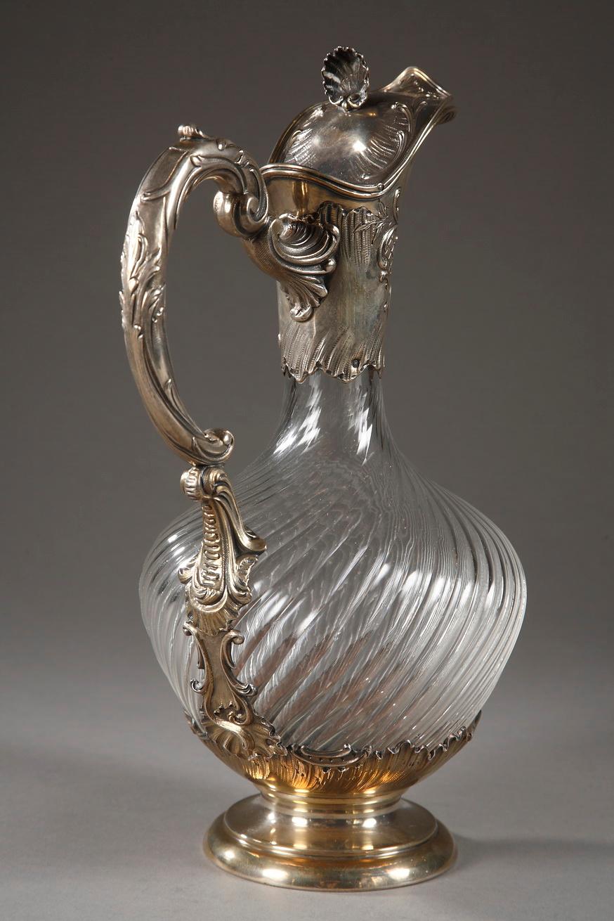 Cut-crystal ewer with twisting patterns and silver mounts. The collar and the base are decorated with stylized waves, diamonds, and foliage, and a shell adorns the top of the pitcher. Its high, C-shaped handle is embellished with acanthus leaves.