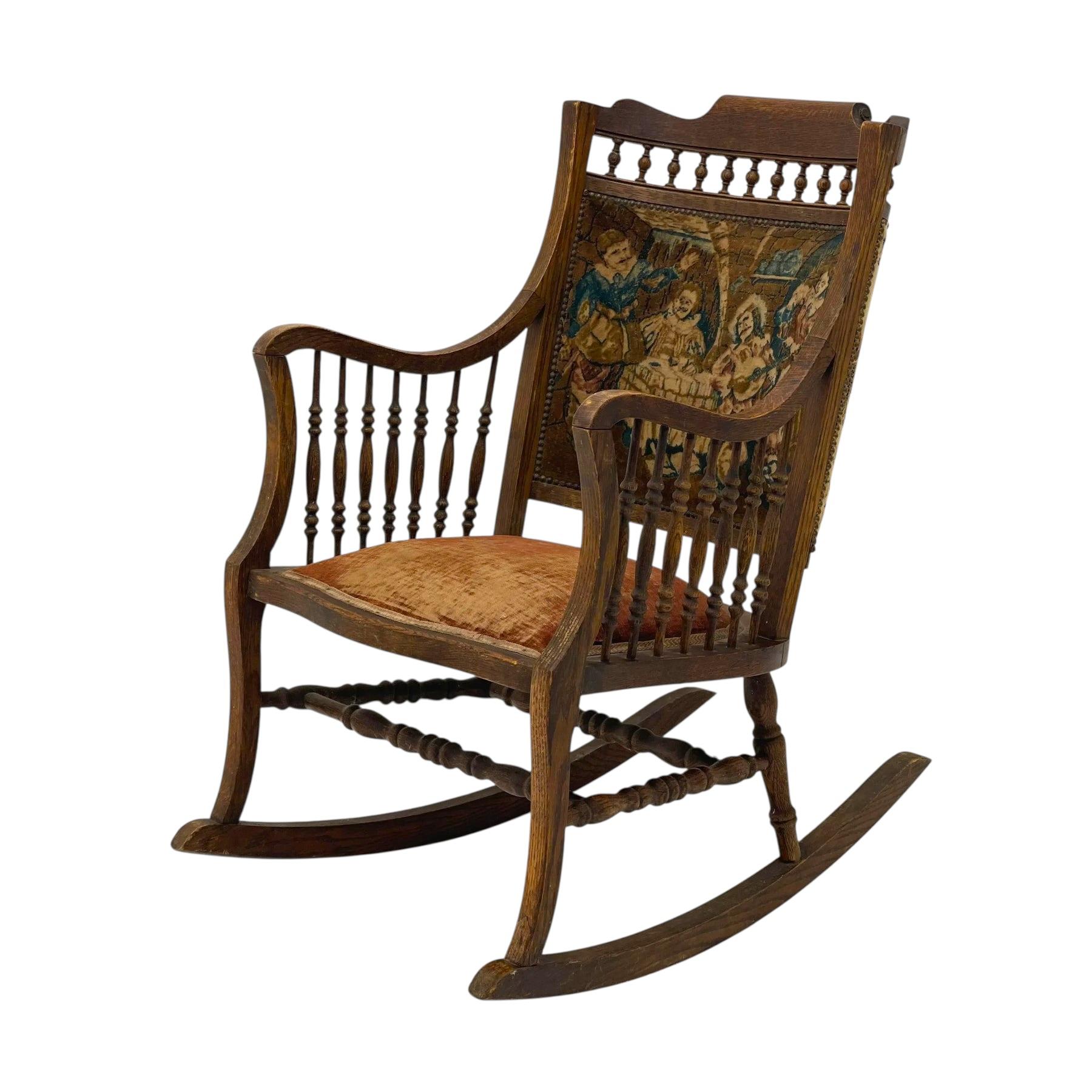 Esta mecedora antigua tiene un armazón de madera con husillos torneados y reposabrazos curvados, de estilo victoriano. El respaldo está tapizado con un tapiz que representa una escena histórica o pastoril, mientras que el asiento está acolchado con