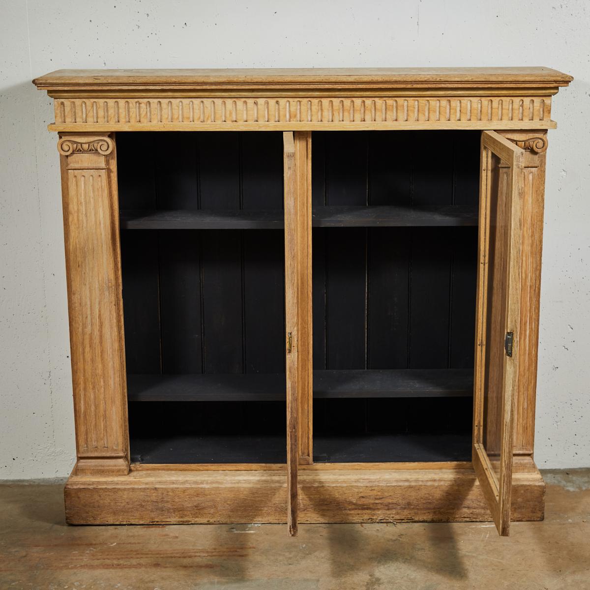 Late 19th-century English bleached oak bookcase with glass doors. Features hand-carved ionic pilaster detailing in natural oak with a stunning blond patina. The interior is painted black and visible through the glass doors. A perfect showcase for