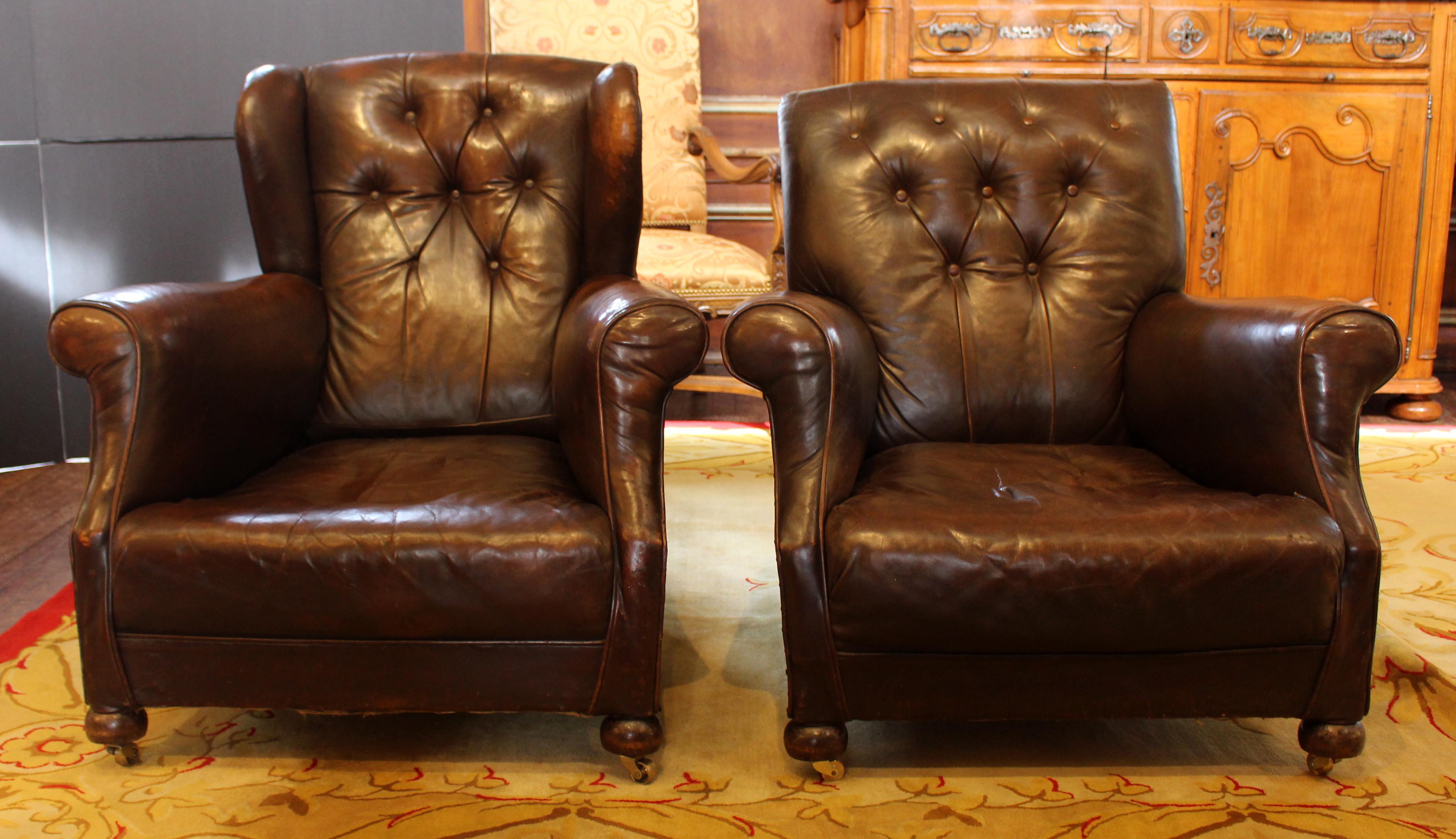 Late 19th century pair of leather club chairs, English. Both with rolled arms, tufted backs, brass studs & bun feet. One with a slight wingback. Original leather. Later casters. One lacks a tufting button. Measures : 39