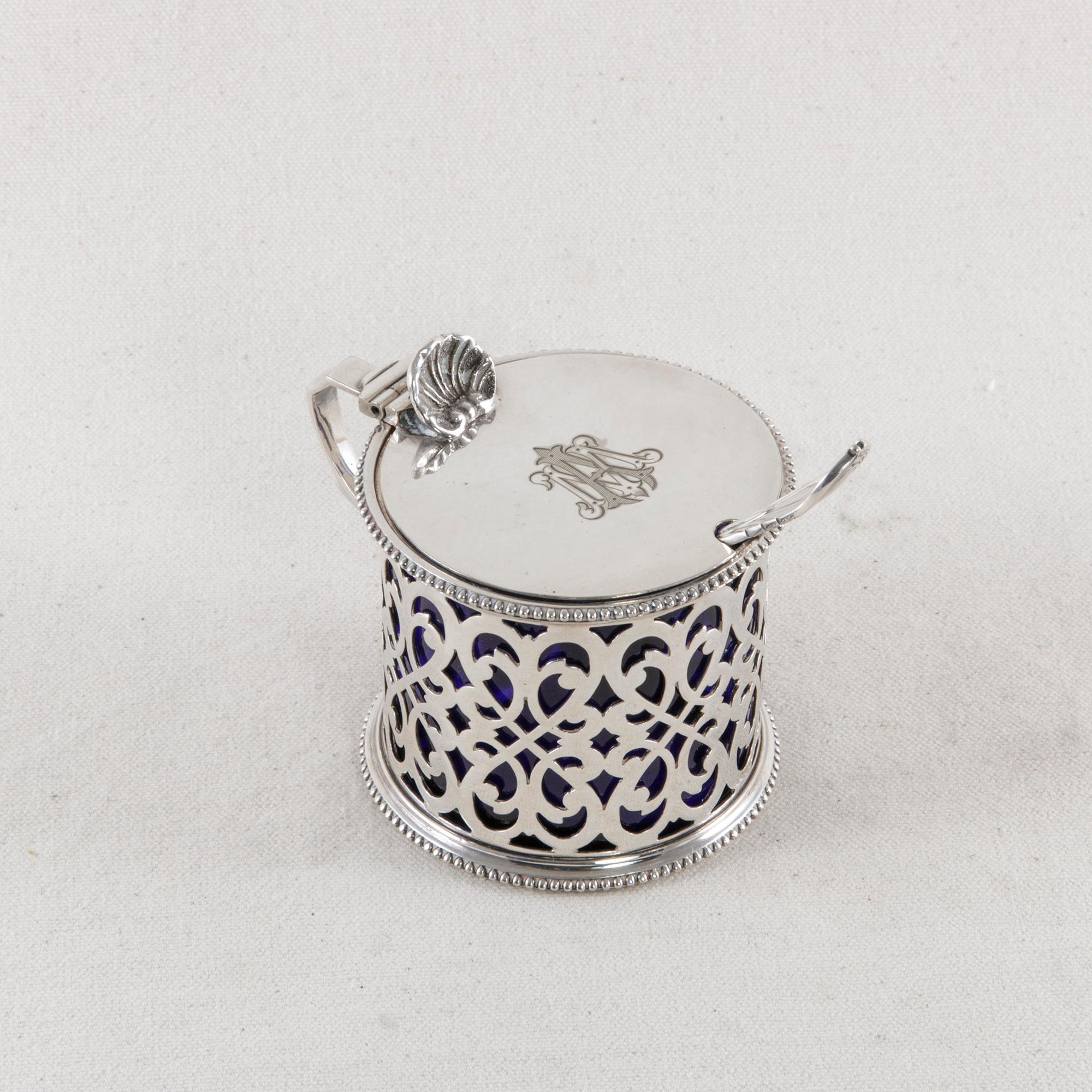 This late 19th century English silver plate mustard pot features the monogram A.M. on its hinged lid. The pierced facade has a scrolling pattern, and beading surrounds the top and bottom. A shell at the top of the handle acts as a thumb lever. On