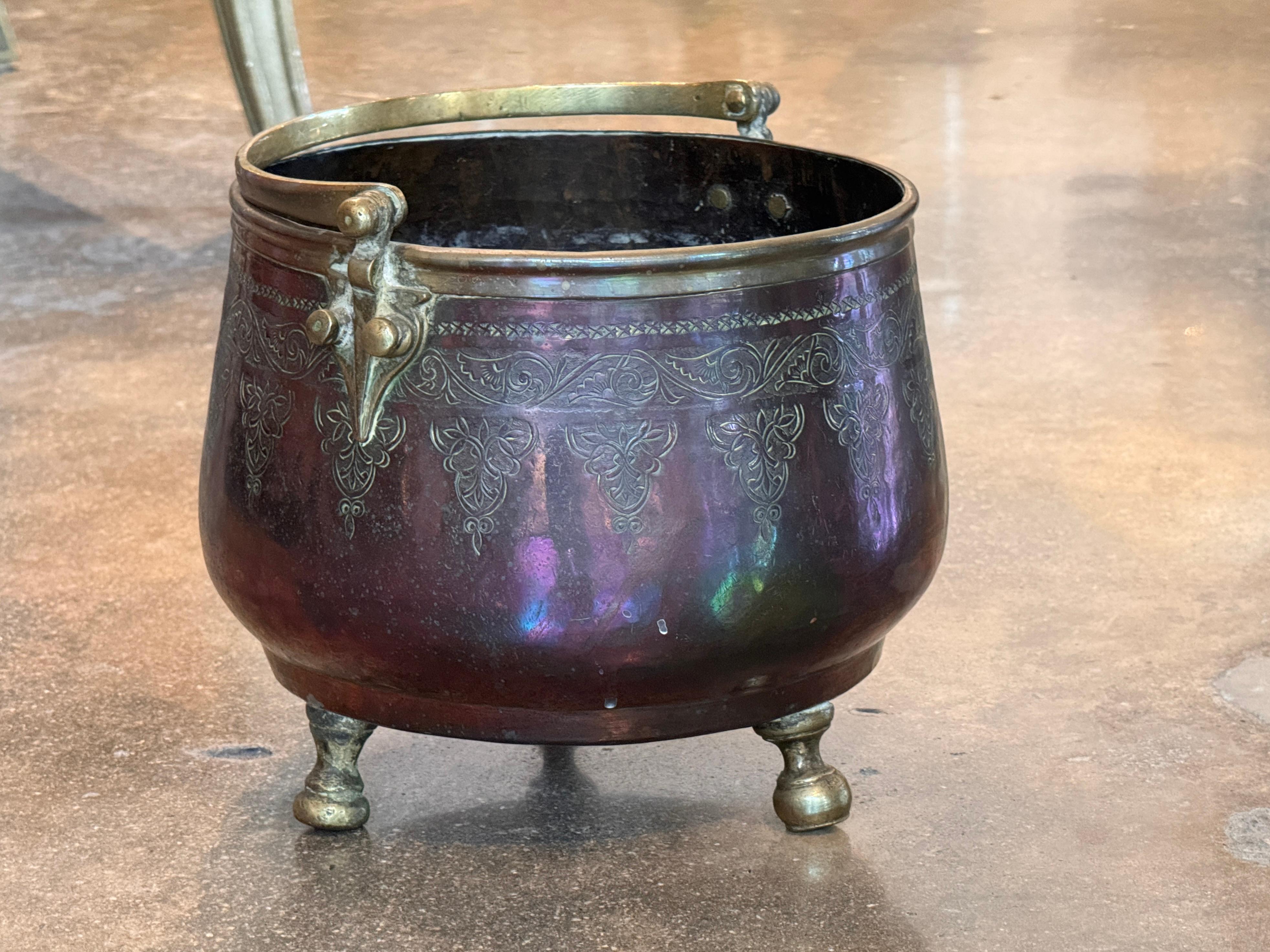 Cubo marroquí con motivos foliados finamente grabados y un cálido acabado patinado. El cuerpo redondeado descansa sobre tres pies torneados de latón y presenta un asa abatible con monturas de latón remachado. La imagen en primer plano destaca los