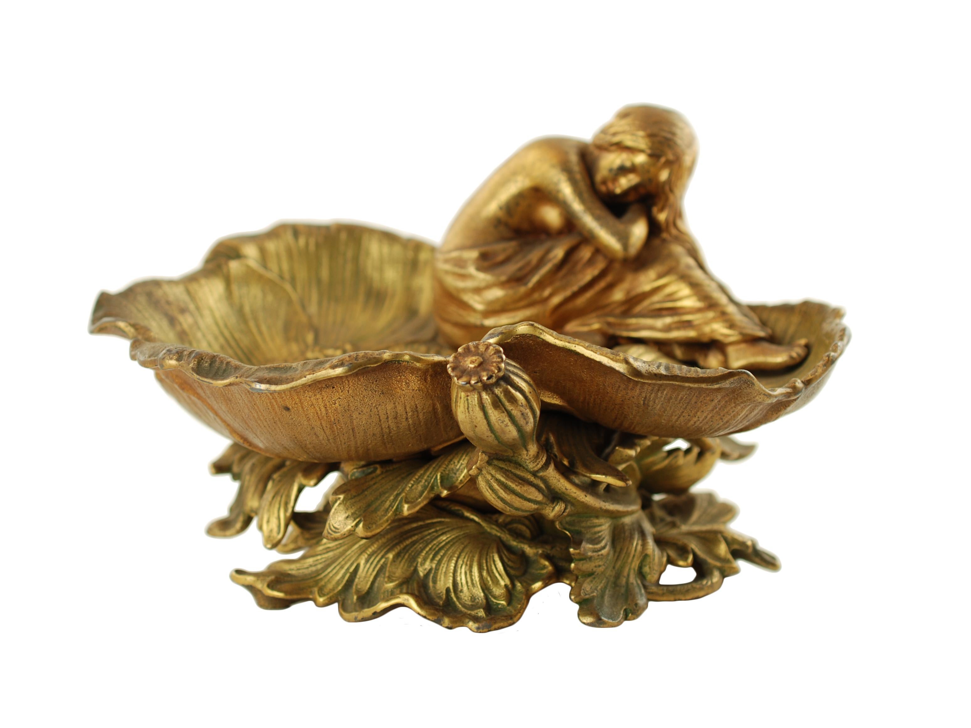 This heavy centerpiece bowl dates to the late 19th century and is composed of cast iron with a gilt finish. The bowl has a shallow basin made up of a series of detailed open poppy blossoms and features a sleeping maiden curled up at one side. The