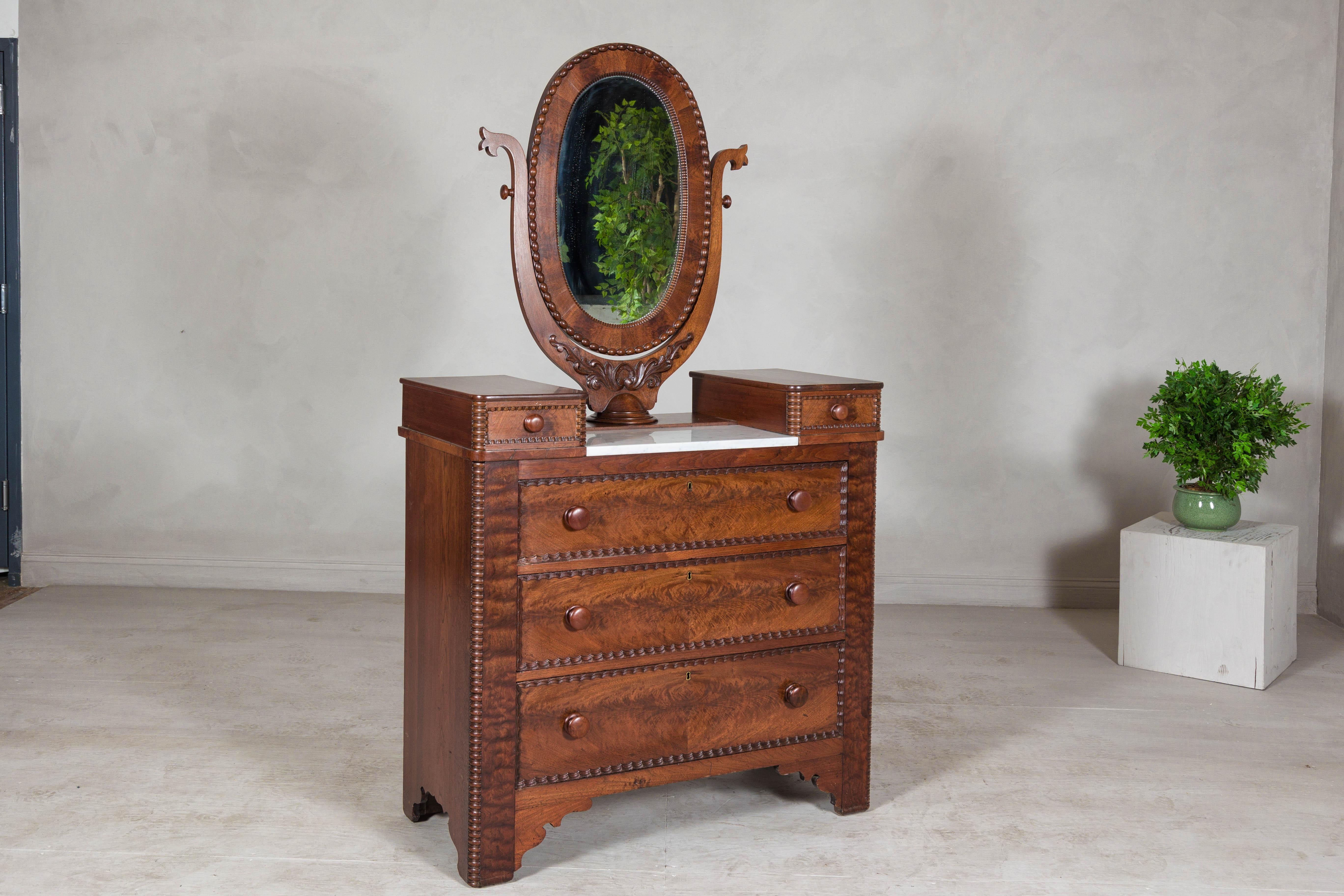 Comò in noce fiammato in stile Mitchell & Rammelsberg della fine del XIX secolo con specchio ovale inclinabile, inserto in marmo bianco e frontali dei cassetti intricatamente intagliati. Questo comò antico incarna l'eleganza dei mobili americani