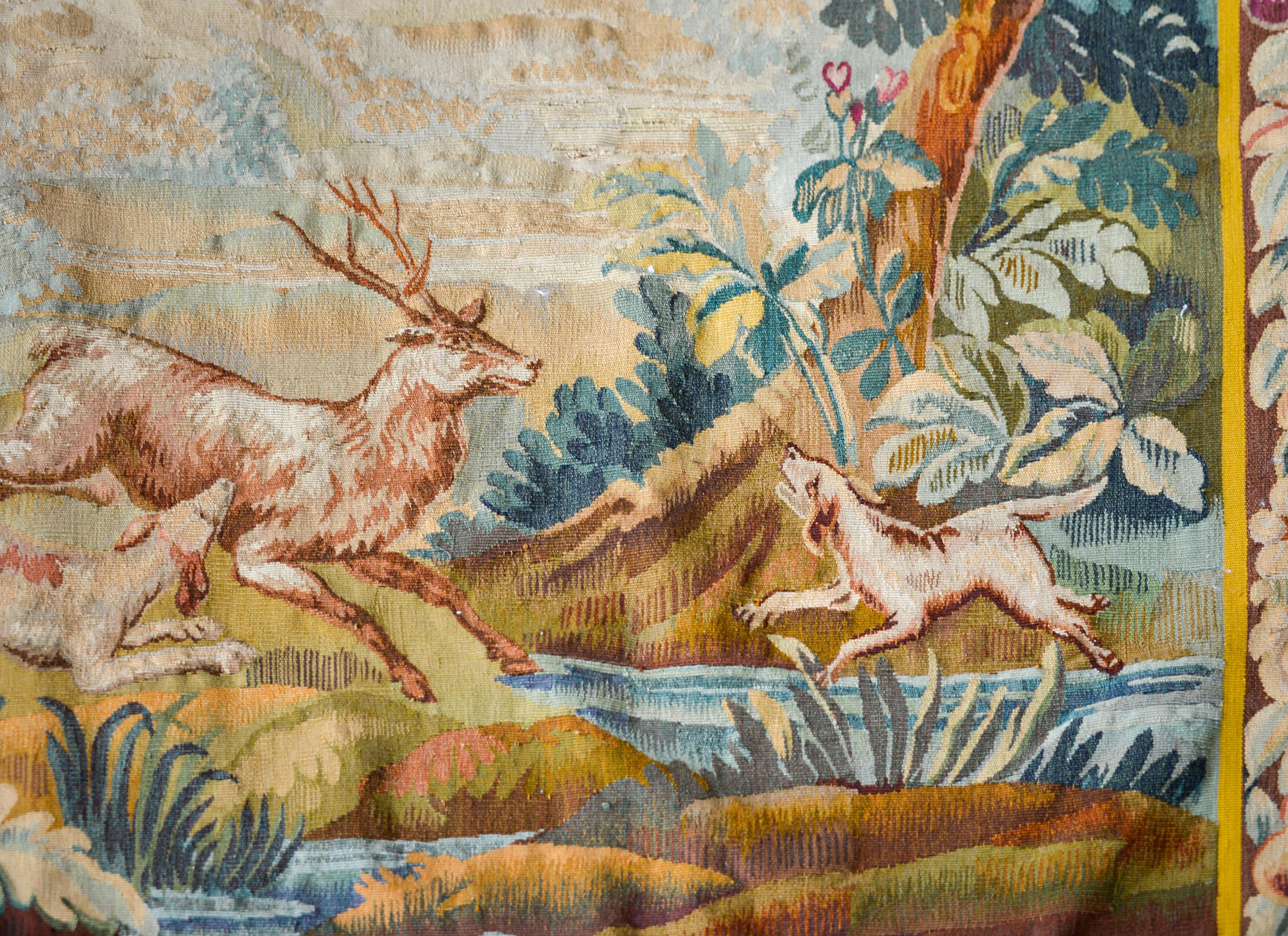 A beautiful late 19th century French Aubusson tapestry depicting a hunt scene with a hunter mounted on horse with two hounds chasing a stag through a forest landscape and a castle in the background. The border with woven with a fruit and floral
