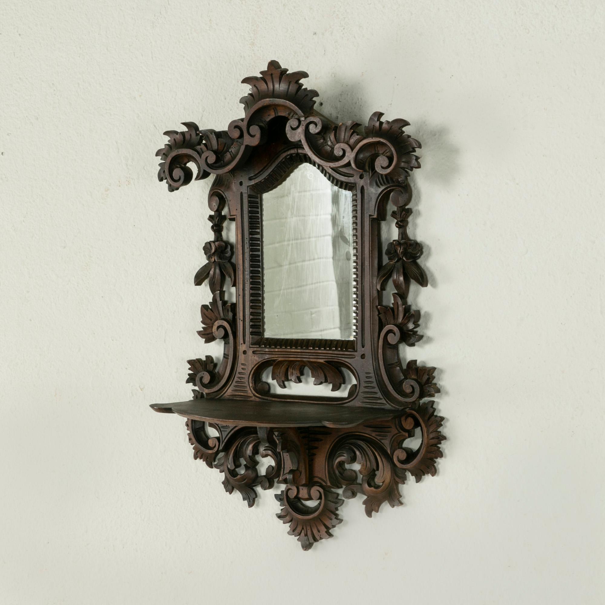 This late nineteenth century French wall mirror with shelf is from the Black Forest region in eastern France. Constructed of hand carved walnut, this piece features a scrolling leaves and floral motif. A shelf below the mirror allows for an object
