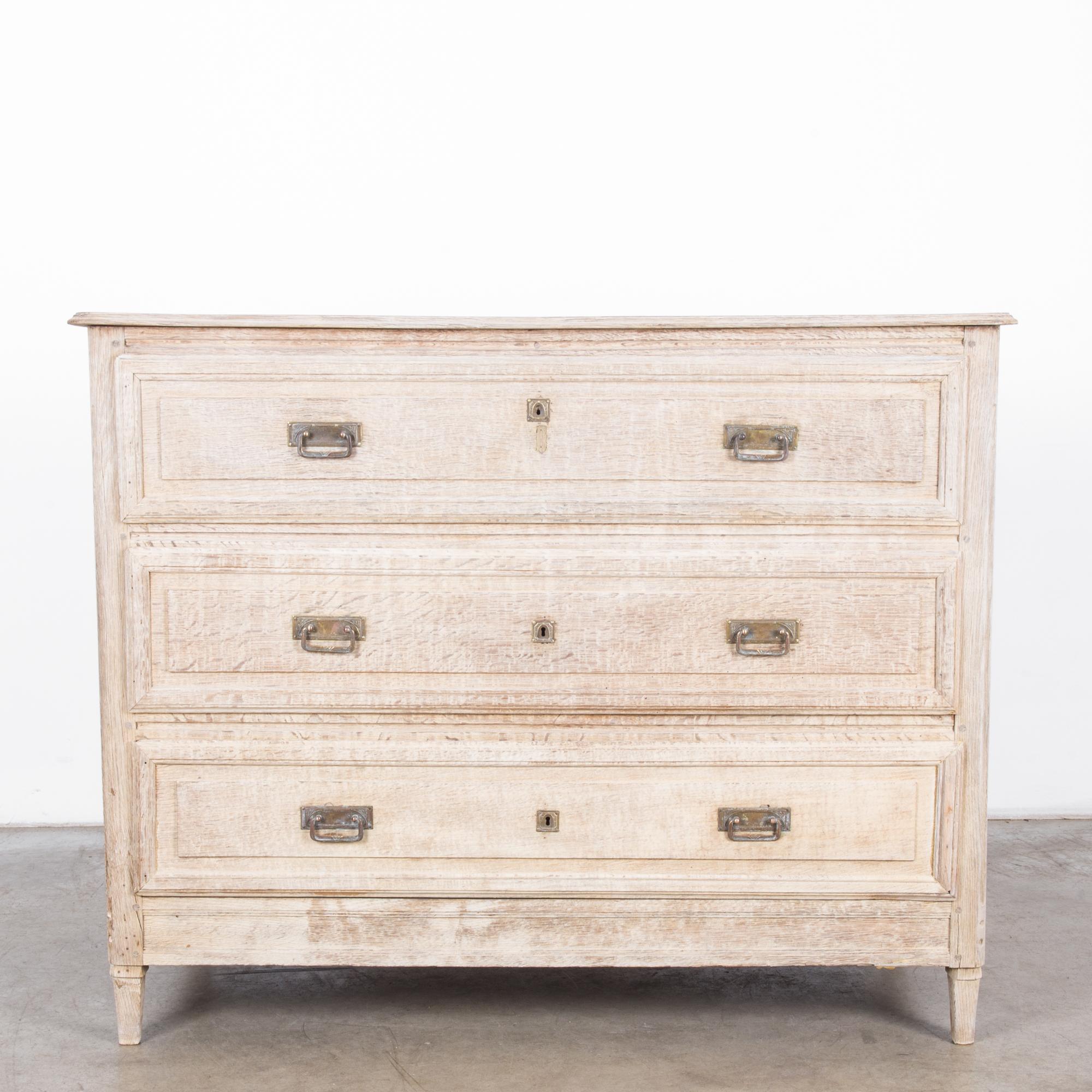 An antique bleached oak chest of drawers from France, circa 1880. The expert craftsmanship of this chest features three wide drawers with plated pulls and decorative keyholes. This impressive piece thrusts itself forward on four tapered legs. Its