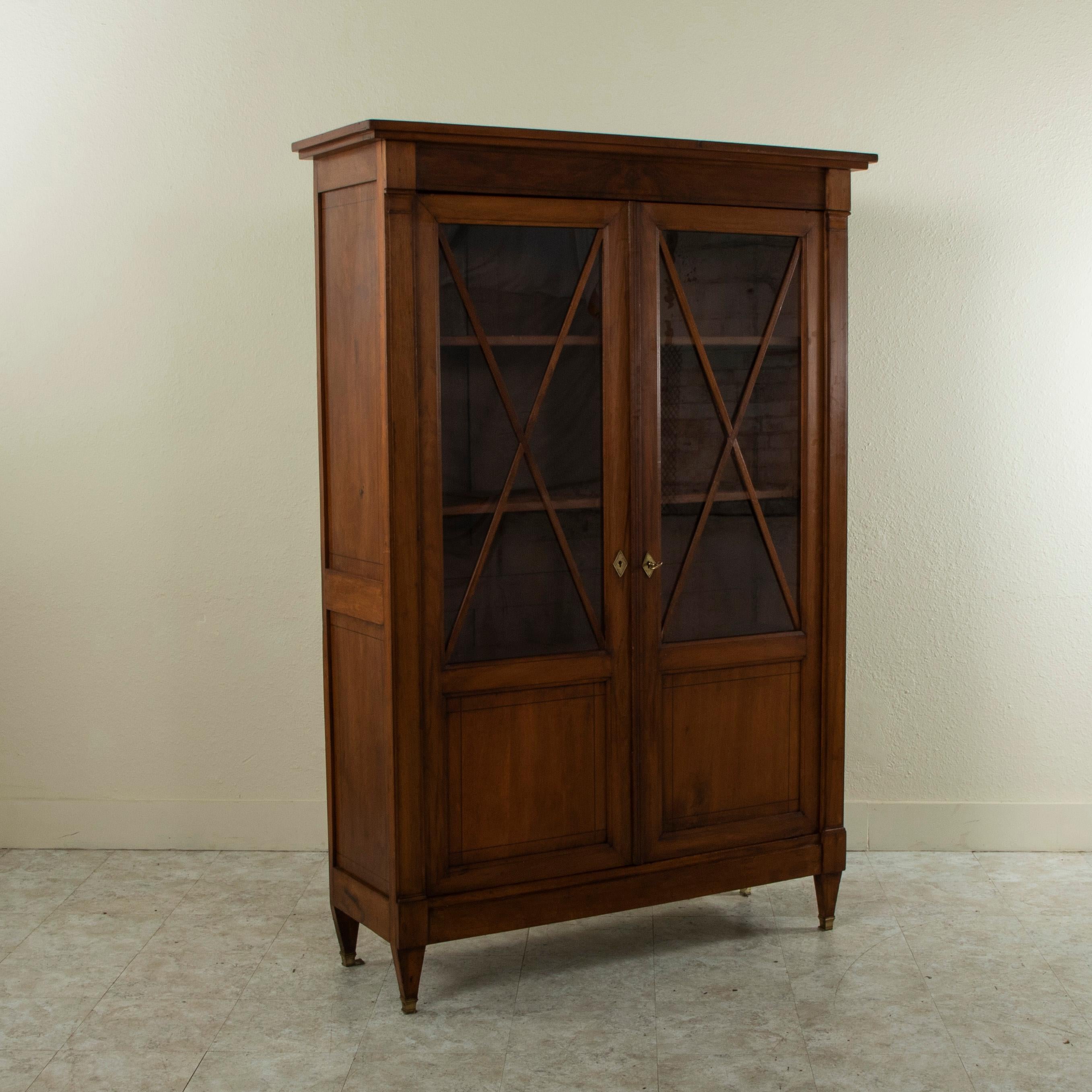 This late nineteenth century French Directoire style bibliotheque or bookcase is constructed of solid walnut and rests on tapered square legs fitted with bronze sabots. Its two glass doors feature crossed lines of walnut over the glass. The doors