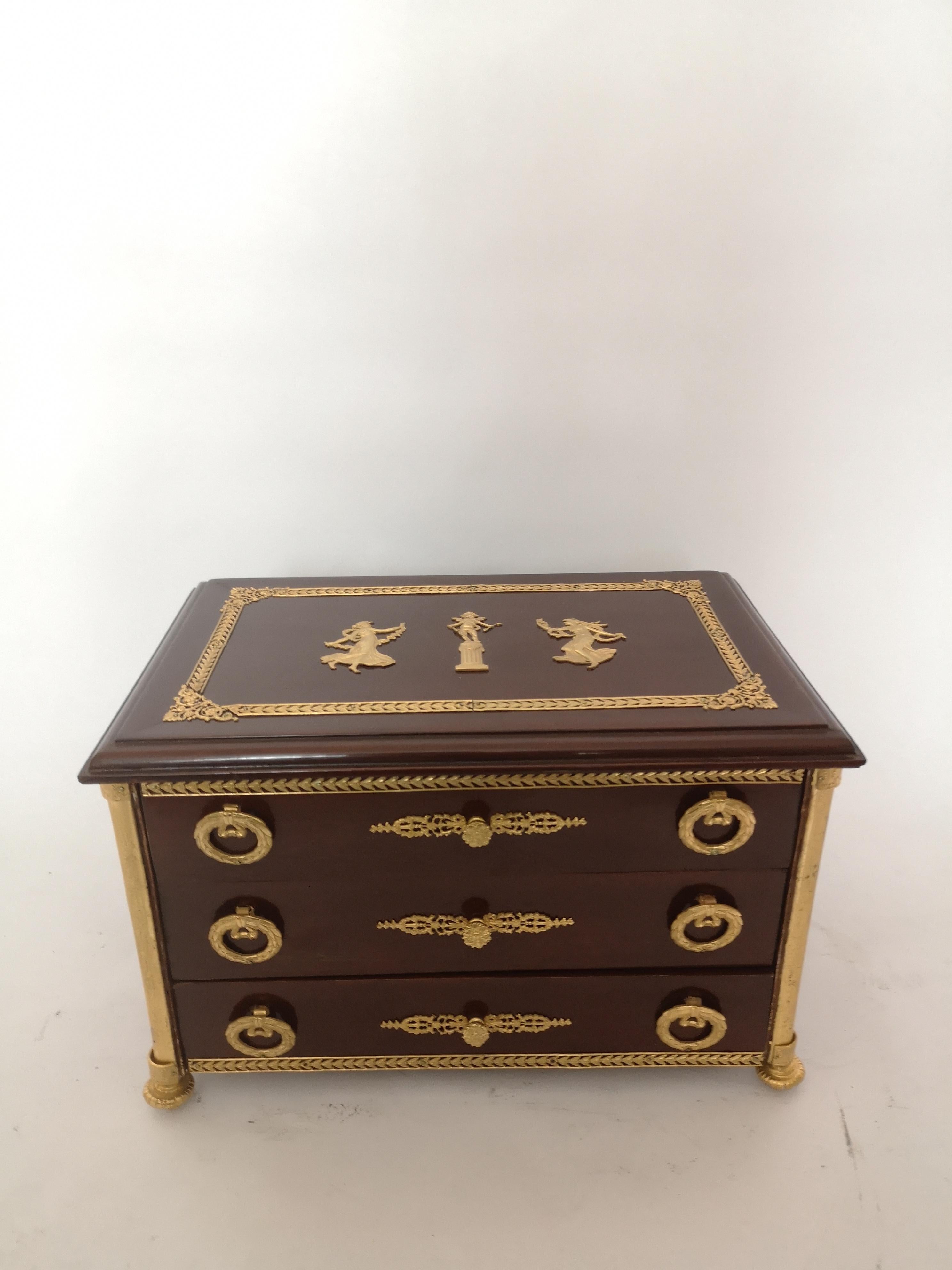 A rare French Empire style miniature chest, made from mahogany with gilt bronze mounts. The top and sides decorated with cherub and classical maiden mounts. The three drawers with wreath handles and a bronze knob, standing on four bronze
