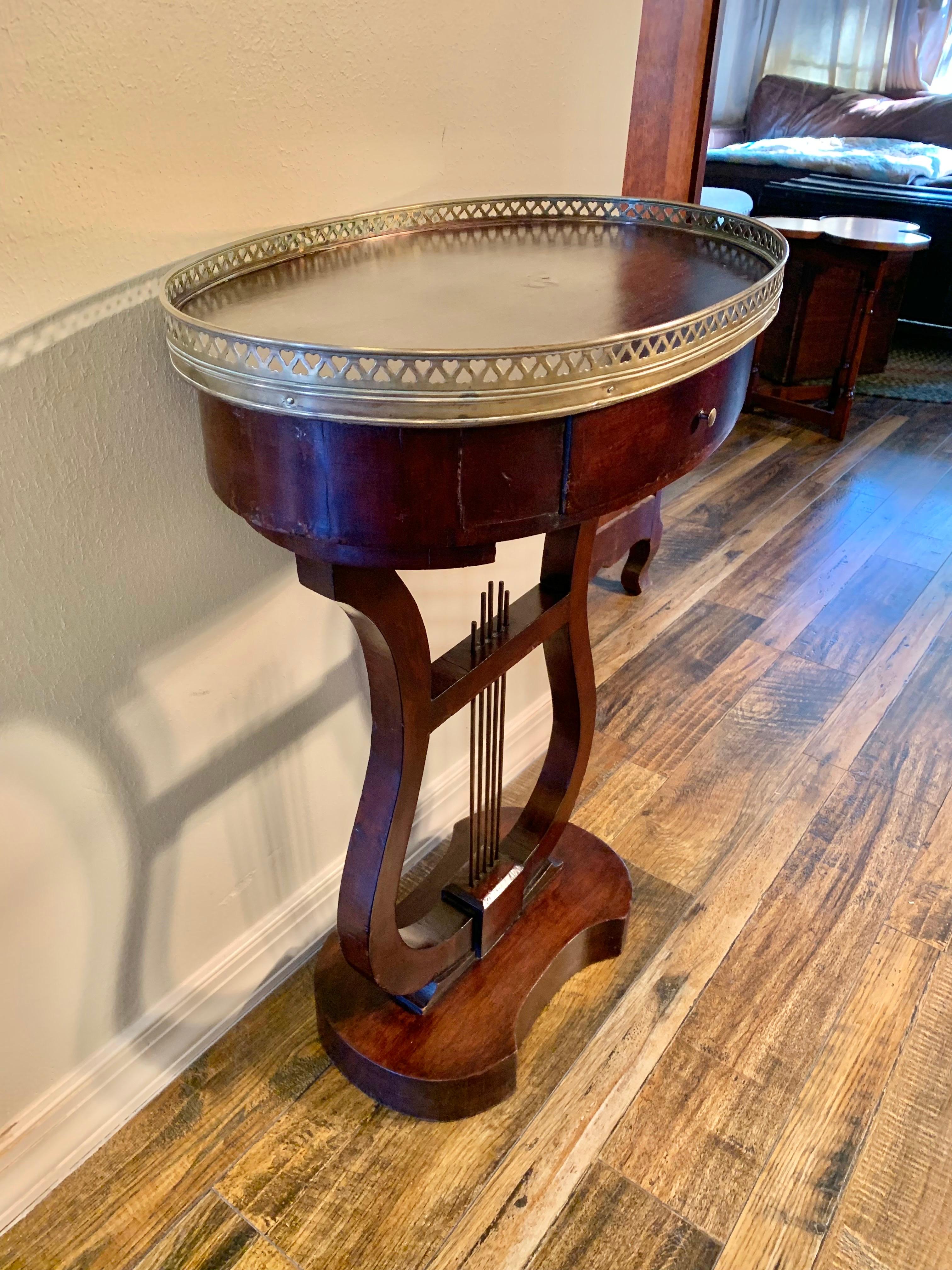 This Late 19th Century French Harp or Lyre Style Side Table is constructed with a wood oval top lined with a pierced brass gallery. The one drawer is adorned with an antique brass knob and the two harp shaped legs rest on a wood base. Perfect for