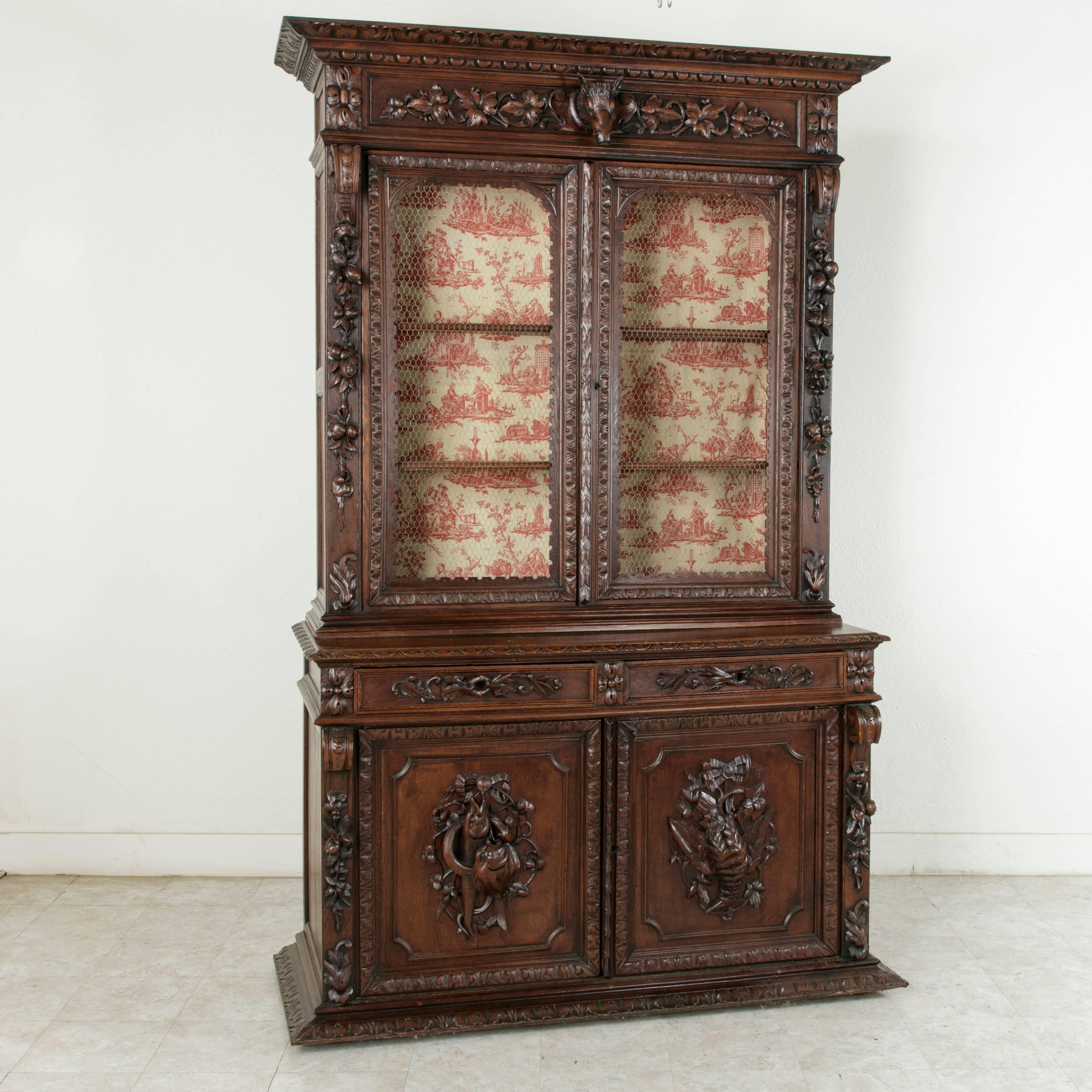 This impressive French oak Henri II style buffet deux corps or bookcase from the late 19th century features detailed hand carvings with hunting and fishing attributes. The upper cabinet displays a wild boar's head at its crown surrounded by grape