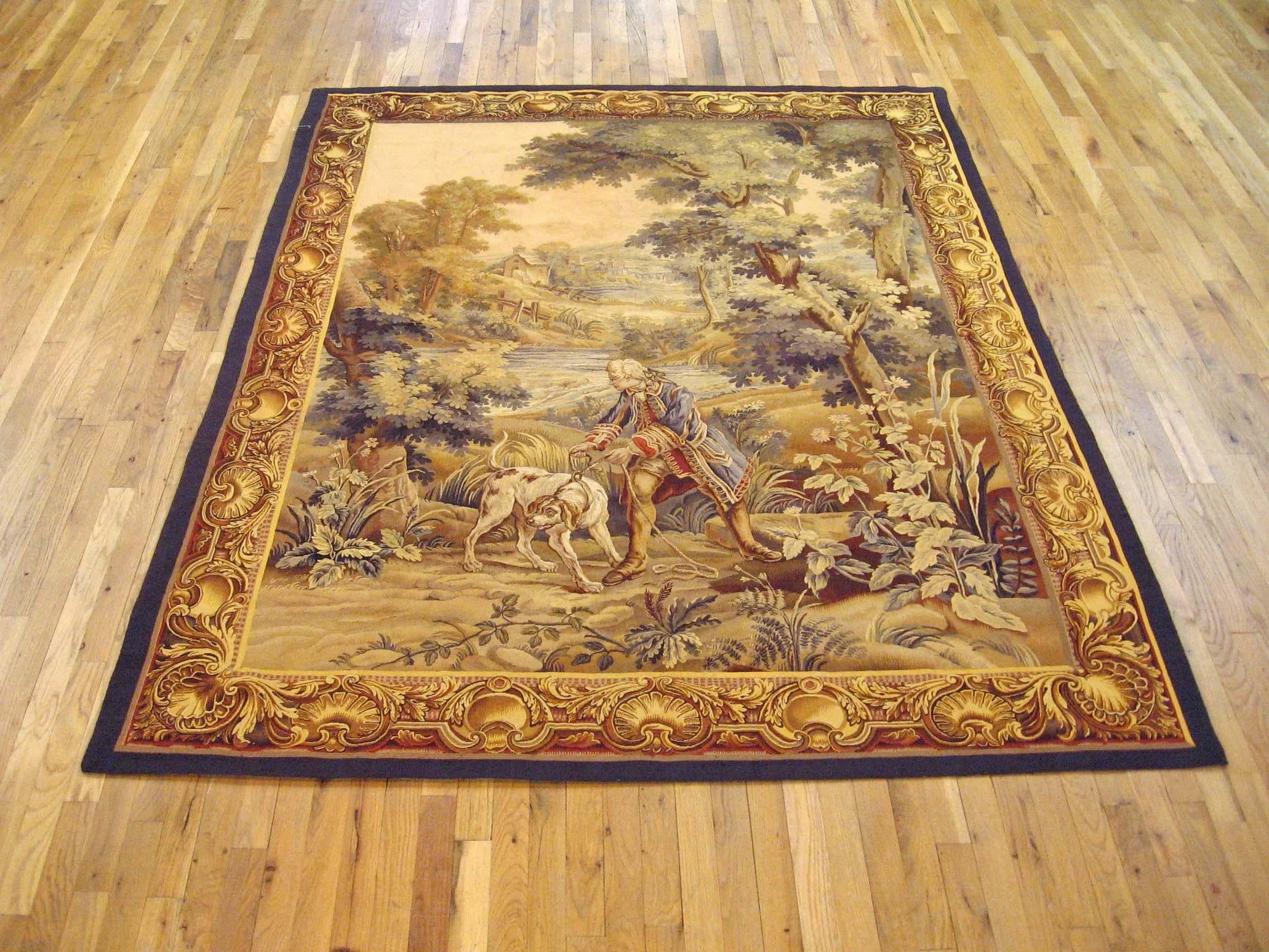 A French hunting tapestry from the late 19th century, envisioning a scene in which a nobleman leads his hound through a verdant area in search of the day’s hunt, with a stately tree at right and a pond and rolling hills in the background. Enclosed