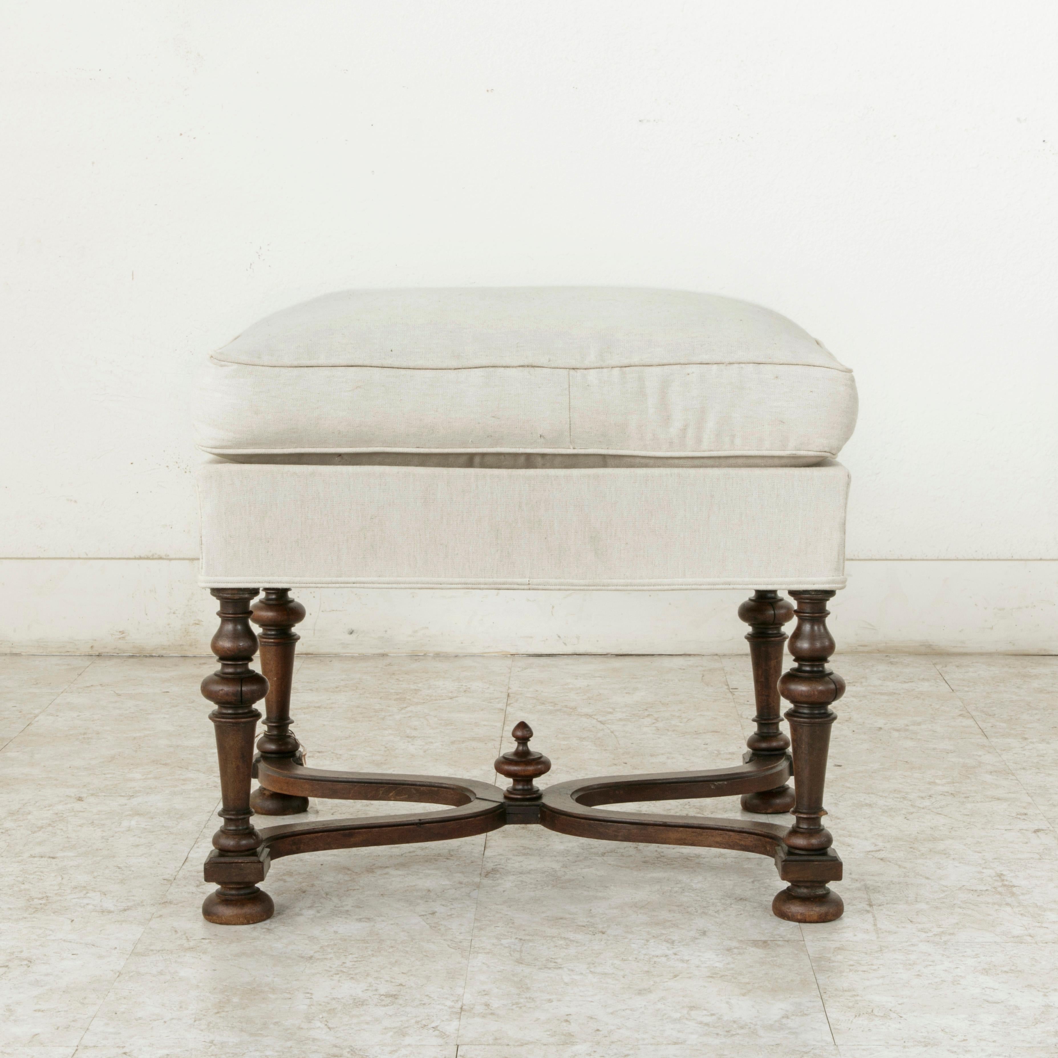 This late 19th French Louis XIII style ottoman is constructed of solid beechwood. Its turned legs are joined by a curved X-stretcher with a central finial. The seat and cushion are upholstered in linen, circa 1890.