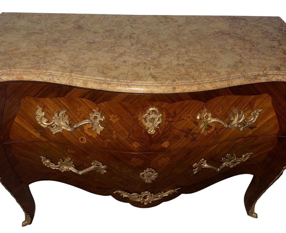 An elegant Louis XV style commode, richly decorated with floral marquetry inlay and mounted with finely cast gilt-bronze ormolu. The serpentine front features two drawers with decorative escutcheons and foliate handles, framed by bombe sides and