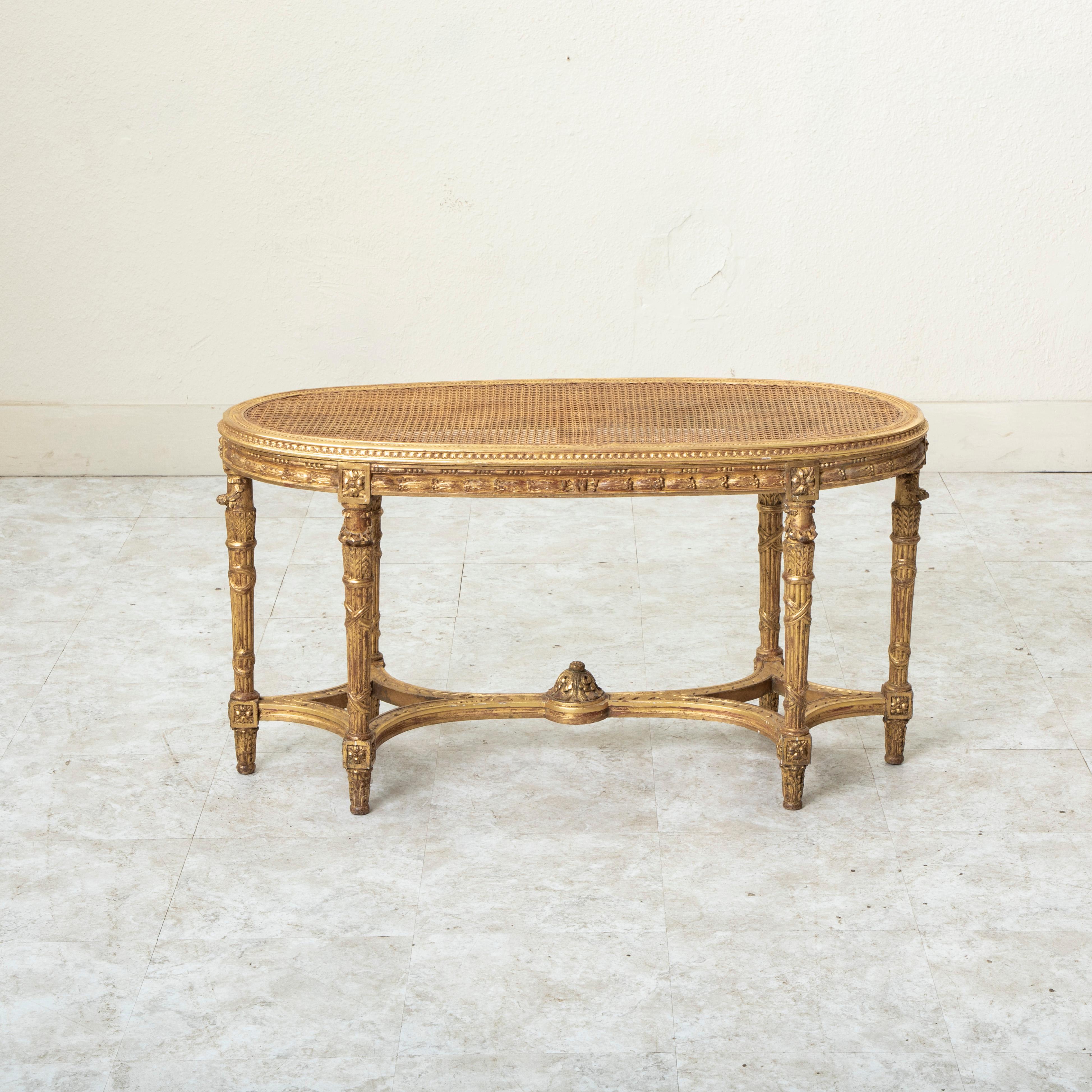 This late nineteenth century French Louis XVI style gilt wood bench features hand carved detailing of beading, crossed ribbons, and rais-de-coeur around its caned seat. Rosettes adorn the die joints with laurels along the apron. The seat rests on