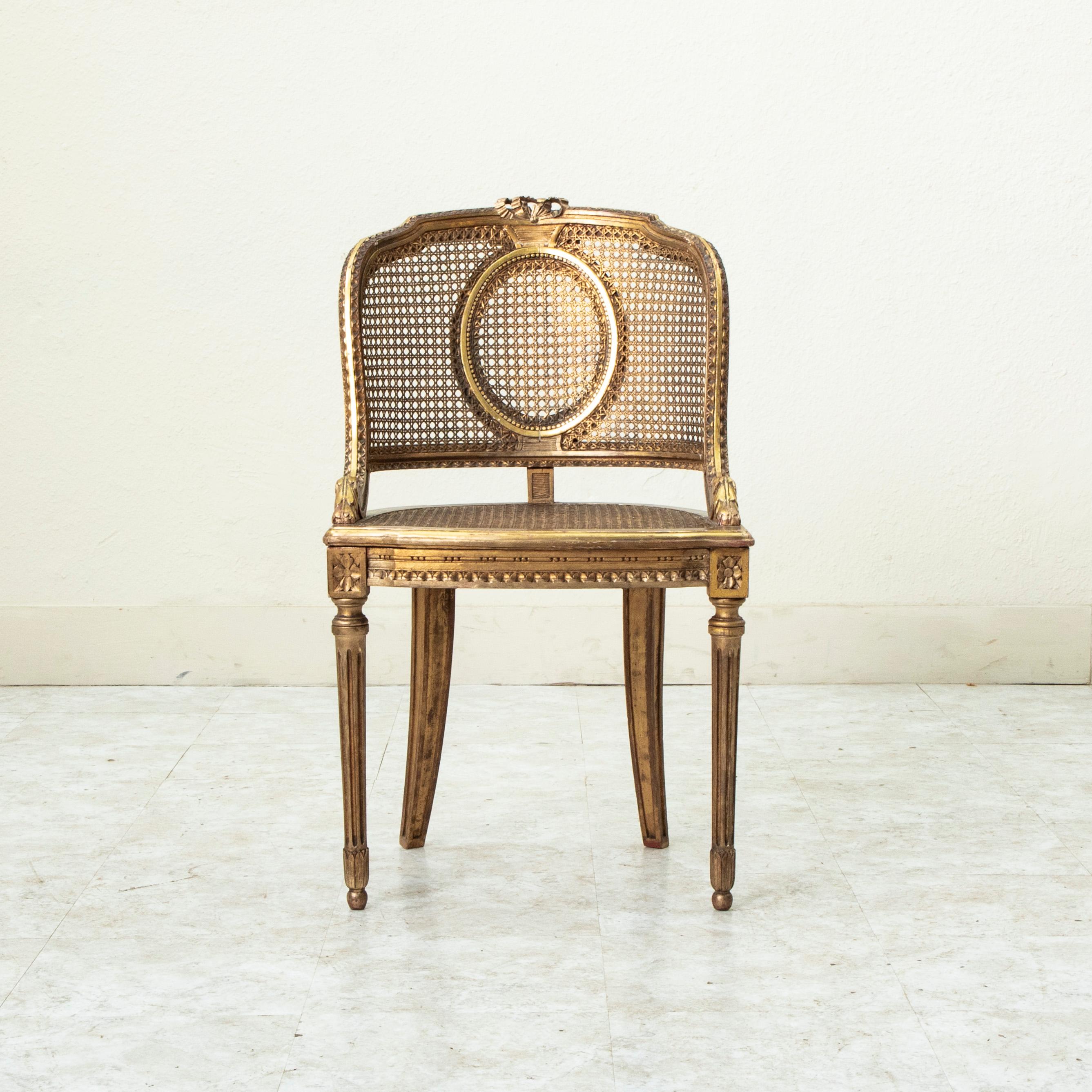 This late nineteenth century Louis XVI style gilt wood bergere or armchair features a caned seat and curved back. The seat back is detailed with a knotted ribbon at the crown as well as a rais-de-coeur pattern and scrolling. Rosettes adorn the die