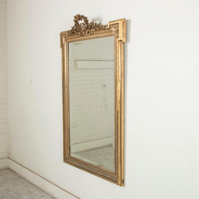 This 19th century hand carved giltwood mirror is a quintessential example of the Louis XVI style. Its 56-inch high frame features Classic Louis XVI motifs of rais-de-coeur, beading, and corner rosettes. A delicately hand carved wreath of roses tied