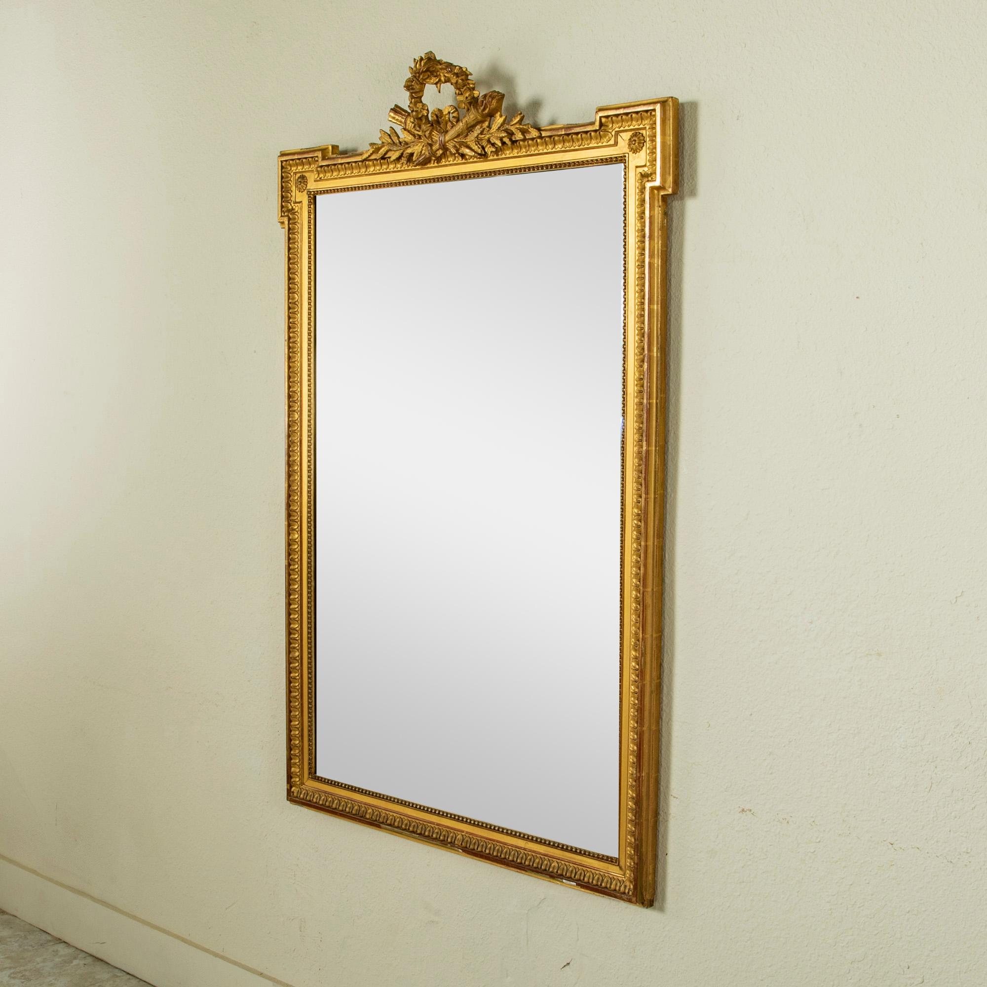 This late nineteenth century hand carved giltwood mirror is a quintessential example of the Louis XVI style. Its 59.5-inch high frame features classic Louis XVI motifs of rais-de-coeur, beading, and corner rosettes. A delicately hand carved wreath