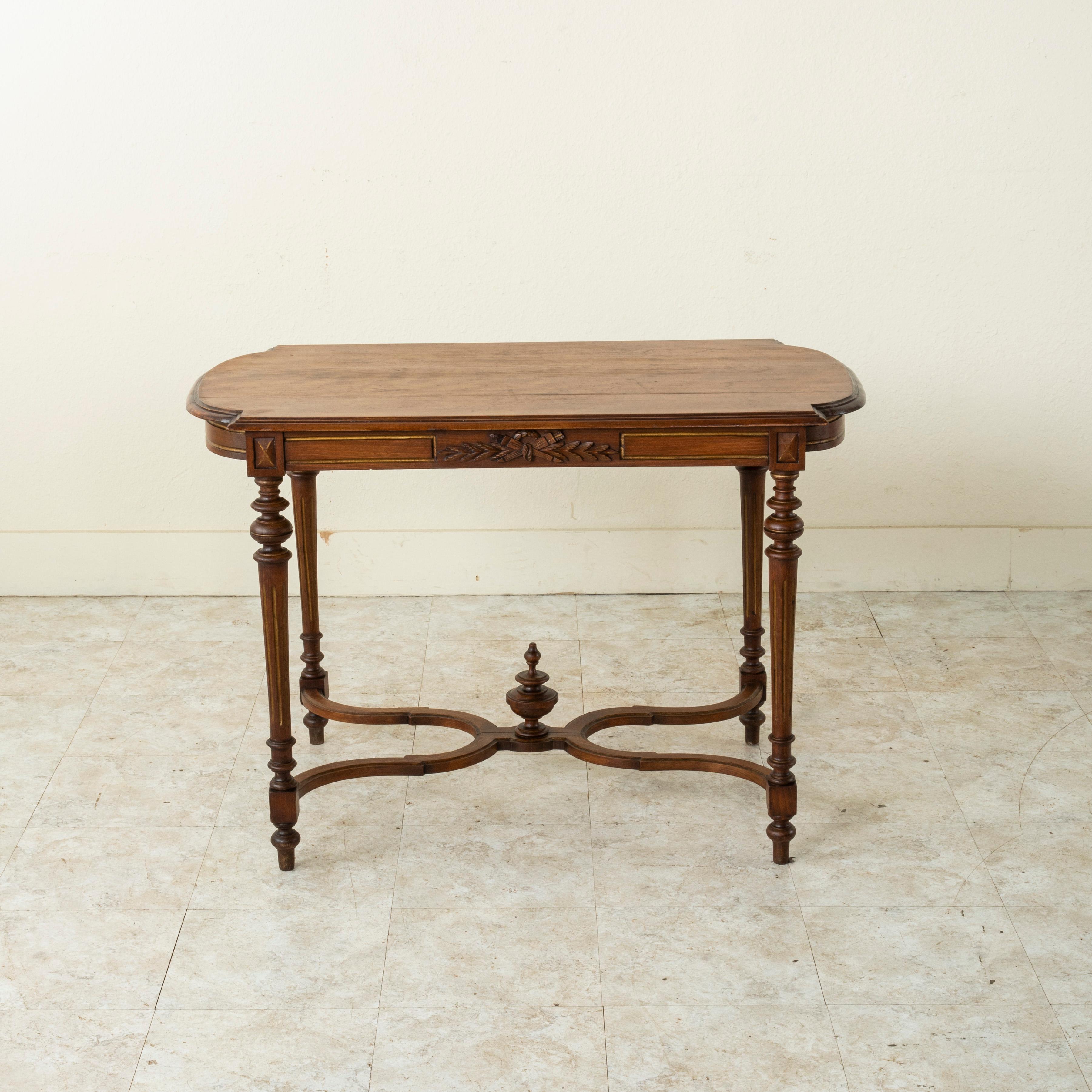 This Louis XVI style walnut writing table from the late 19th century features a hand carved element of a classic crossed torch and quiver with a crown of laurels. Its beveled top rests above an apron detailed with gilded inset lines. Tapered fluted