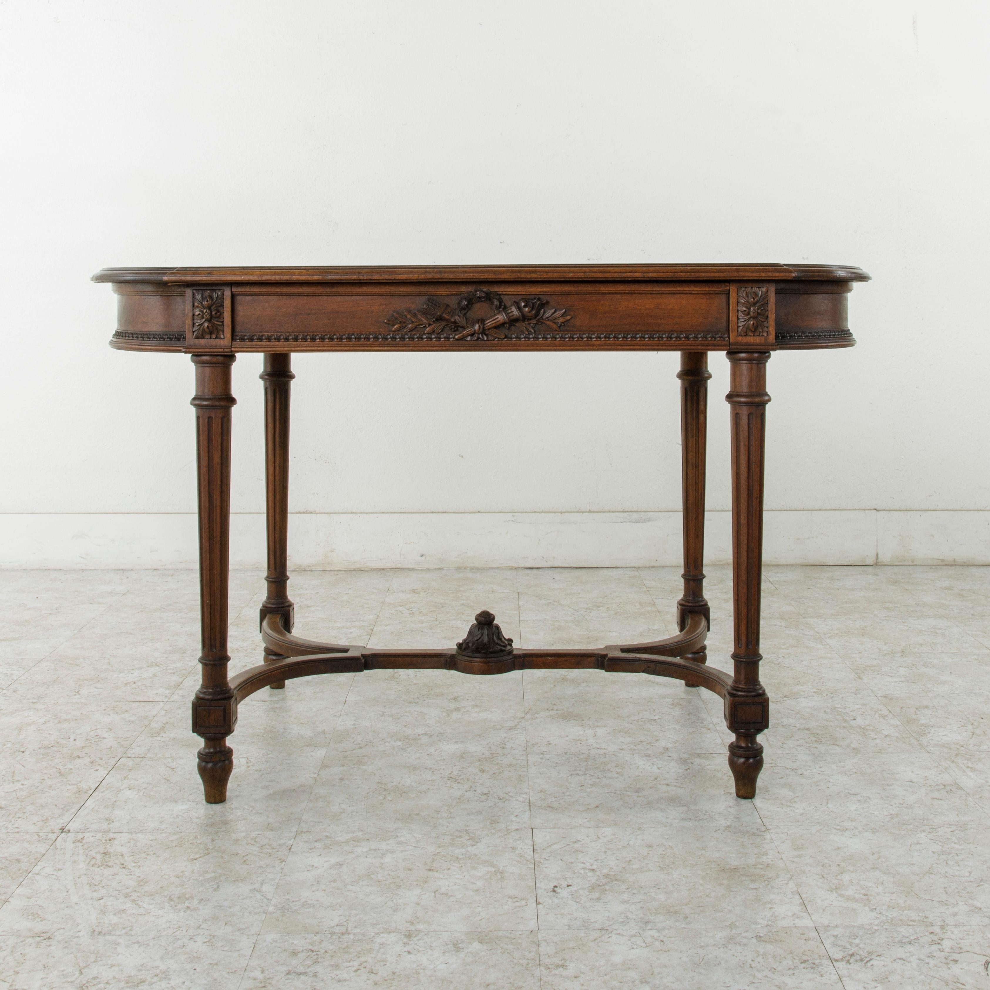 This Louis XVI style walnut writing table or desk from the late 19th century features hand carved detailing of a Classic crossed torch and quiver with laurels and a wreath of roses on two sides. Its beveled top is of oval form, and the apron below