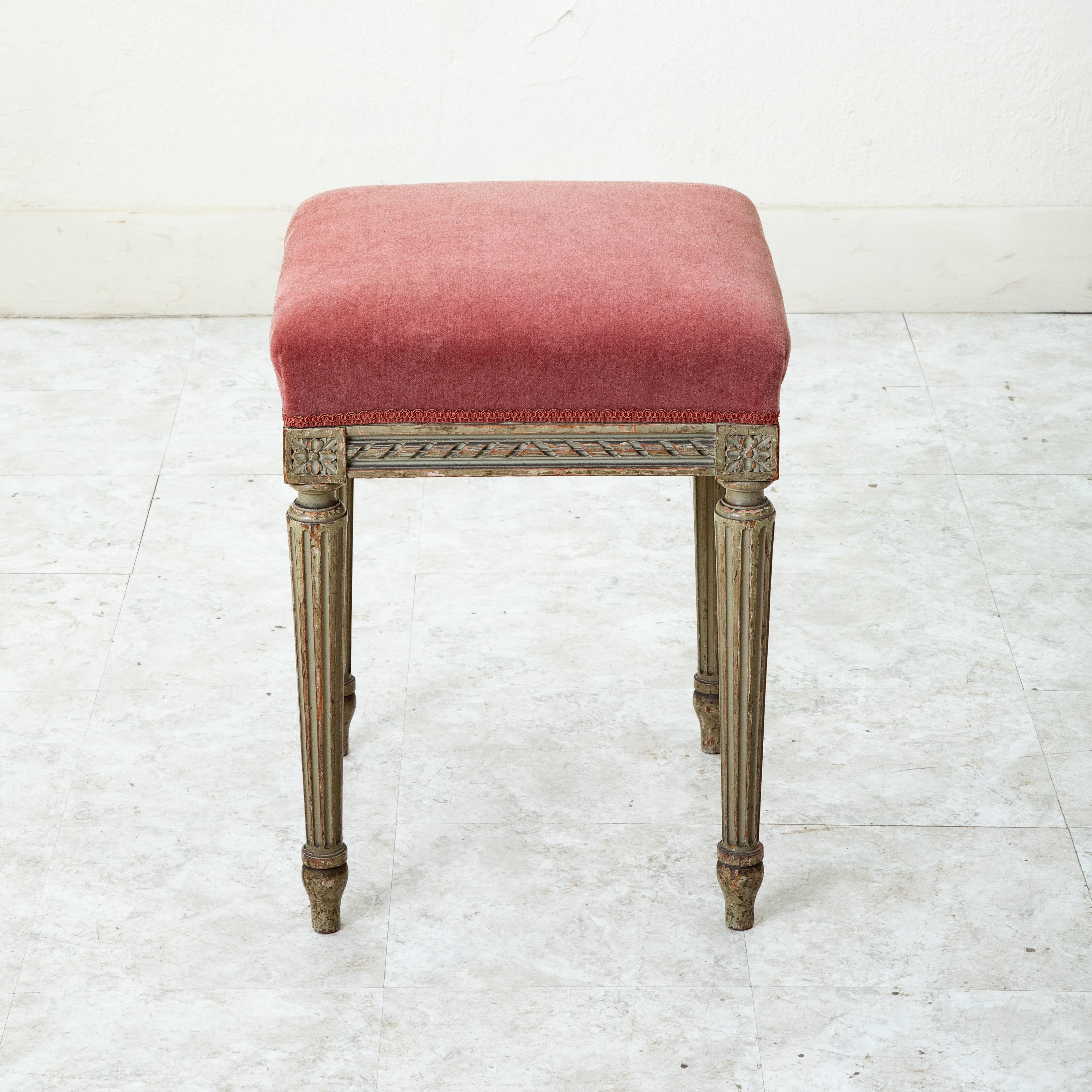 This late nineteenth century French Louis XVI style hand carved and painted vanity stool, bench, or banquette features classic carved rosettes on the die joints above its tapered fluted legs as well as a twisted ribbon motif. Upholstered in pink