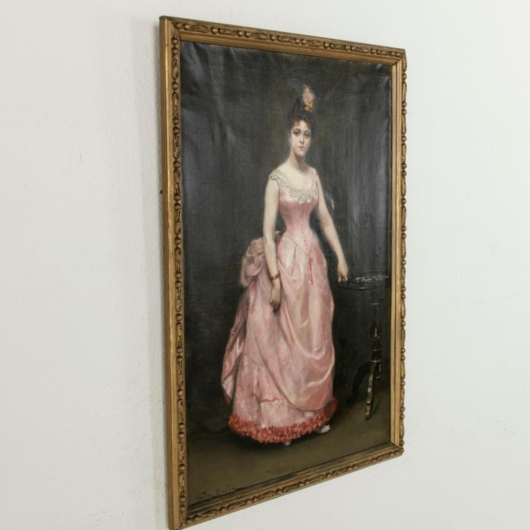 This large scale late 19th century French Napoleon III period oil on canvas features a portrait of a fine lady elegantly dressed in a pink evening gown trimmed in lace. Her hair is coiffed in a chignon and is accented with a ribbon and feather.
