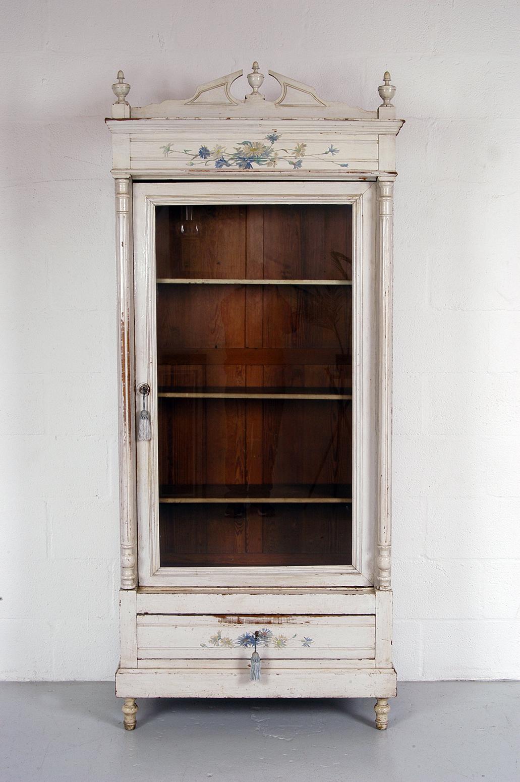 Wonderful French painted pitch pine linen press with three adjustable shelves behind a single glazed door and above a lockable drawer. The top has a removable decorative frieze with three finials. 
The pale grey paint is not original to the piece,