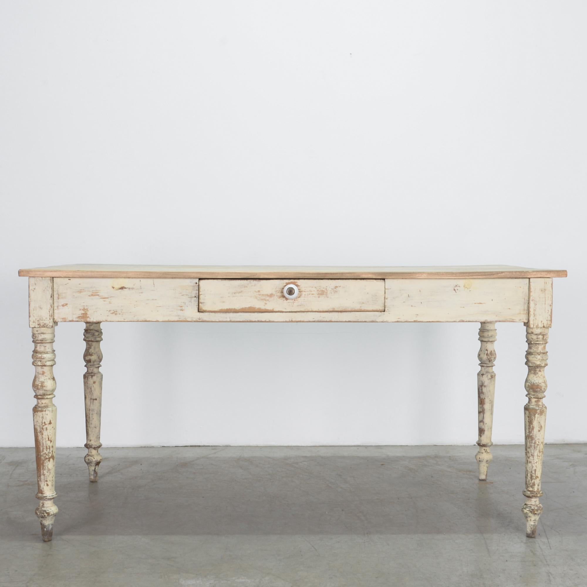 A classical influenced French provincial table with center drawer and porcelain knob. A worn patina with traces of paint and original stained finish, visible through layers gives a casual and rustic effect.