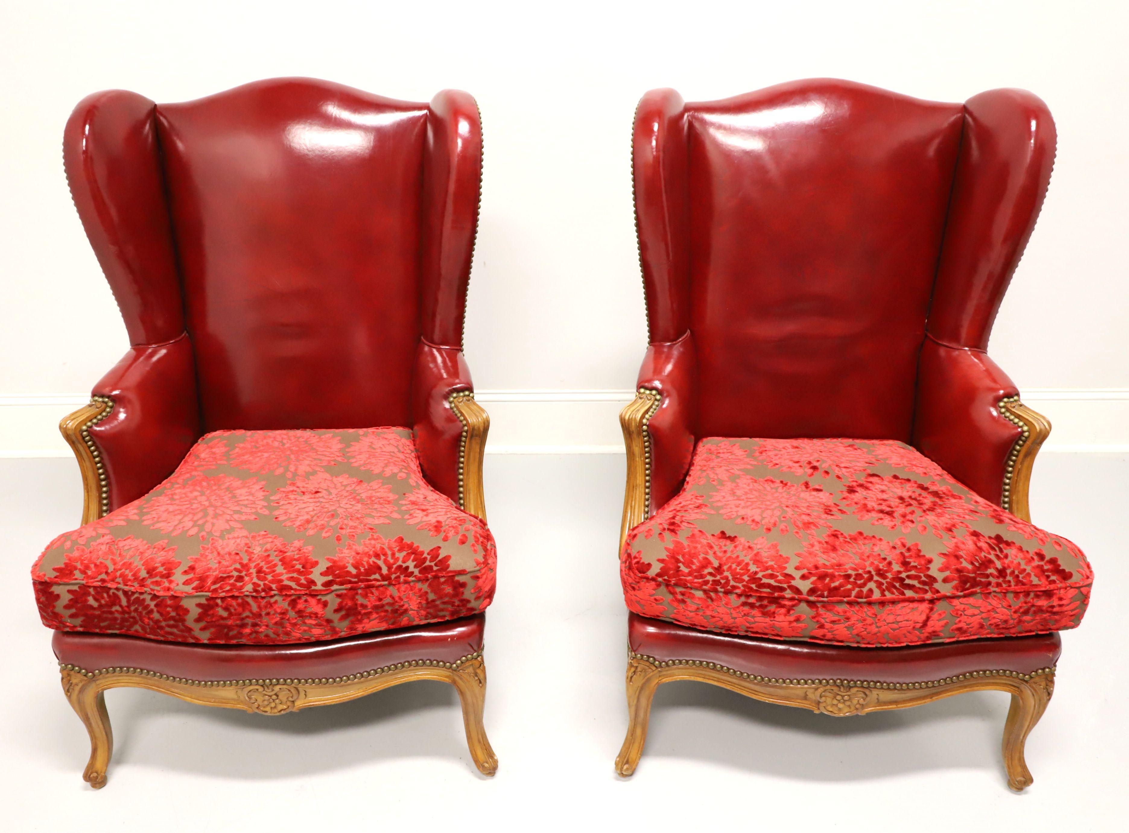 A pair of antique French Provincial Louis XV style wing back chairs, unbranded. Red leather upholstered with crushed velvet brocade fabric seat cushion, brass nailhead trim, nutwood frame with curved carved arms, carved apron, carved knees, curved