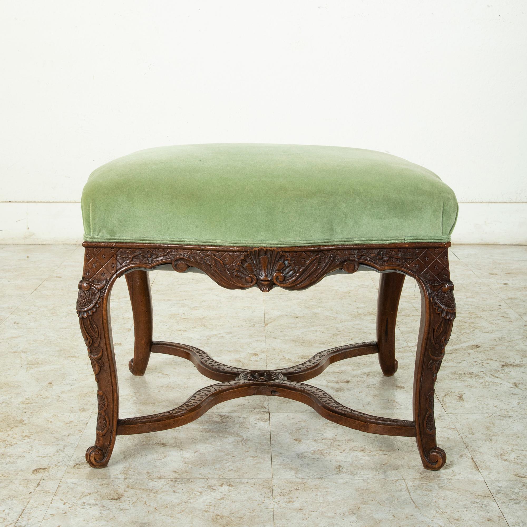 This late 19th century French Regency style oak ottoman or vanity stool features hand carved details of shells, acanthus leaves, and a diamond pattern. The seat rests on cabriolet legs joined by an X stretcher that provides additional stability. The