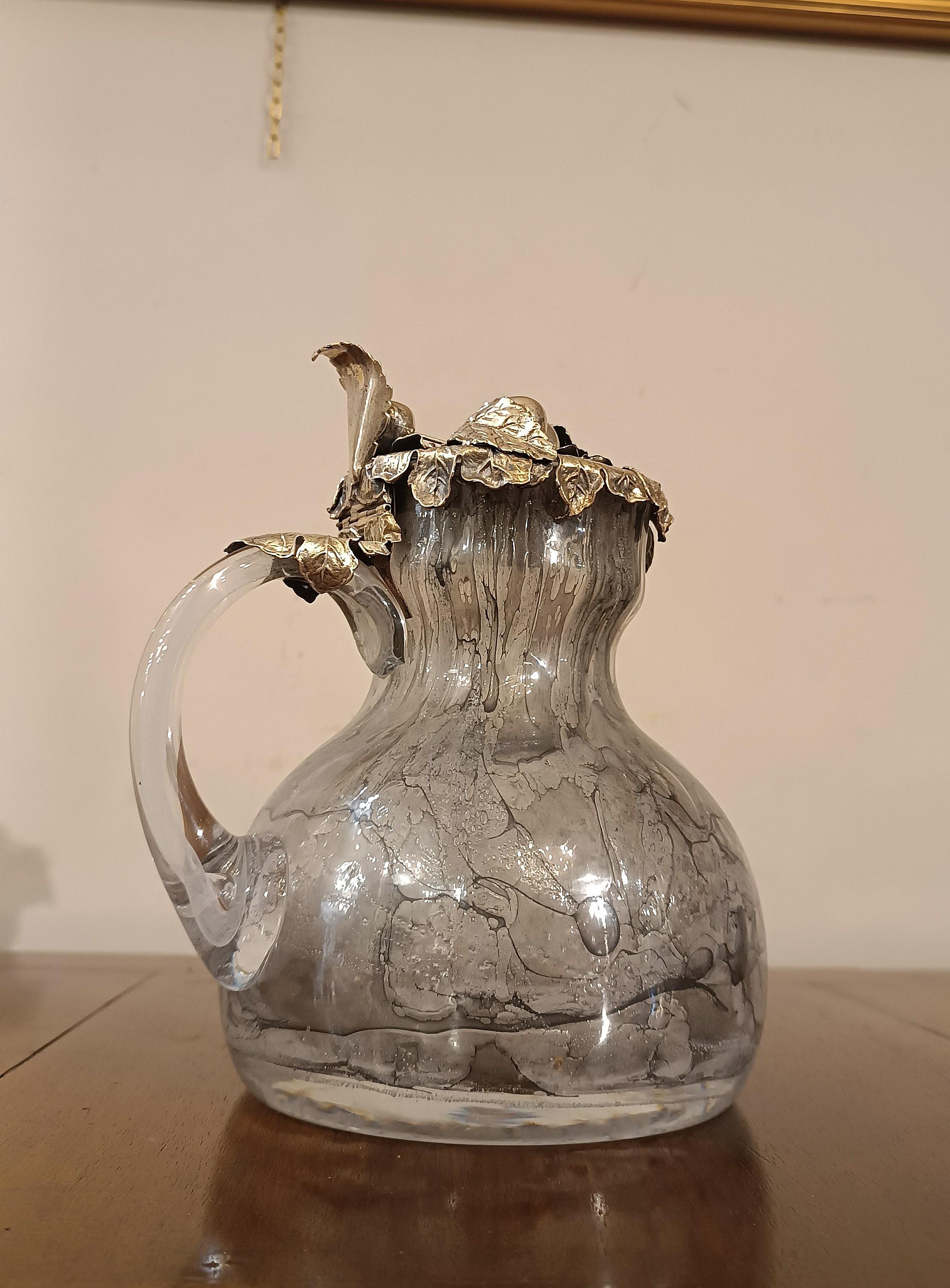 Elegant jug in smoked, colorless and gray glass. This refined jug has a circular body, is equipped with a handle and a silver lid with the 800 nomenclature. The real peculiarity of this jug is the lid, finely decorated with leaf and fruit