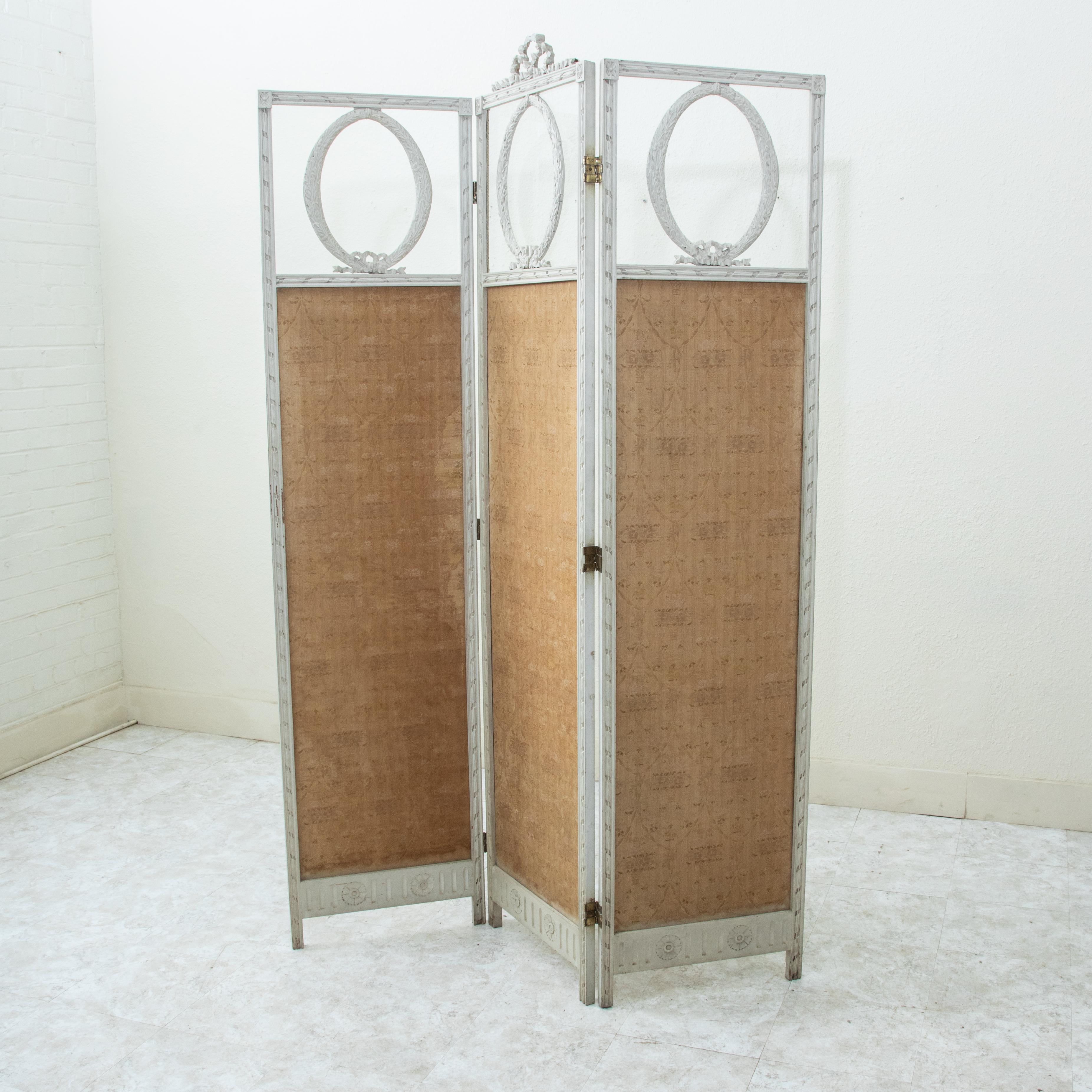 This late nineteenth century French Louis XVI style painted white folding screen or partition is divided into thirds with an upper glass section in each part. A hand carved wreath is inset into the glass pane and a central knotted ribbon crowns the