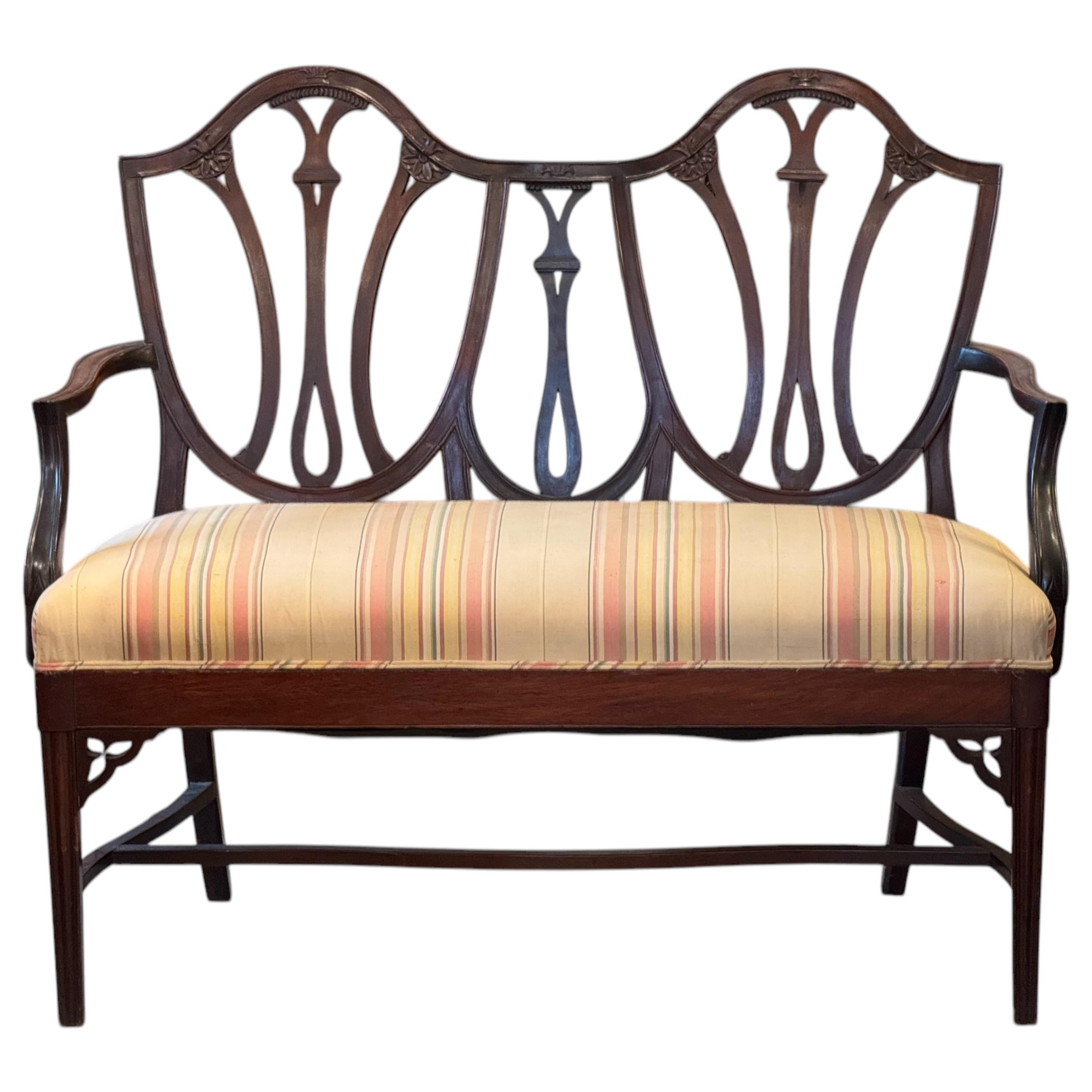Elegant mahogany settee in the Hepplewhite style, featuring a shield-back design with carved floral and fan motifs, gently curved arms, and a stretcher base for added strength. The frame showcases refined neoclassical lines, typical of the late