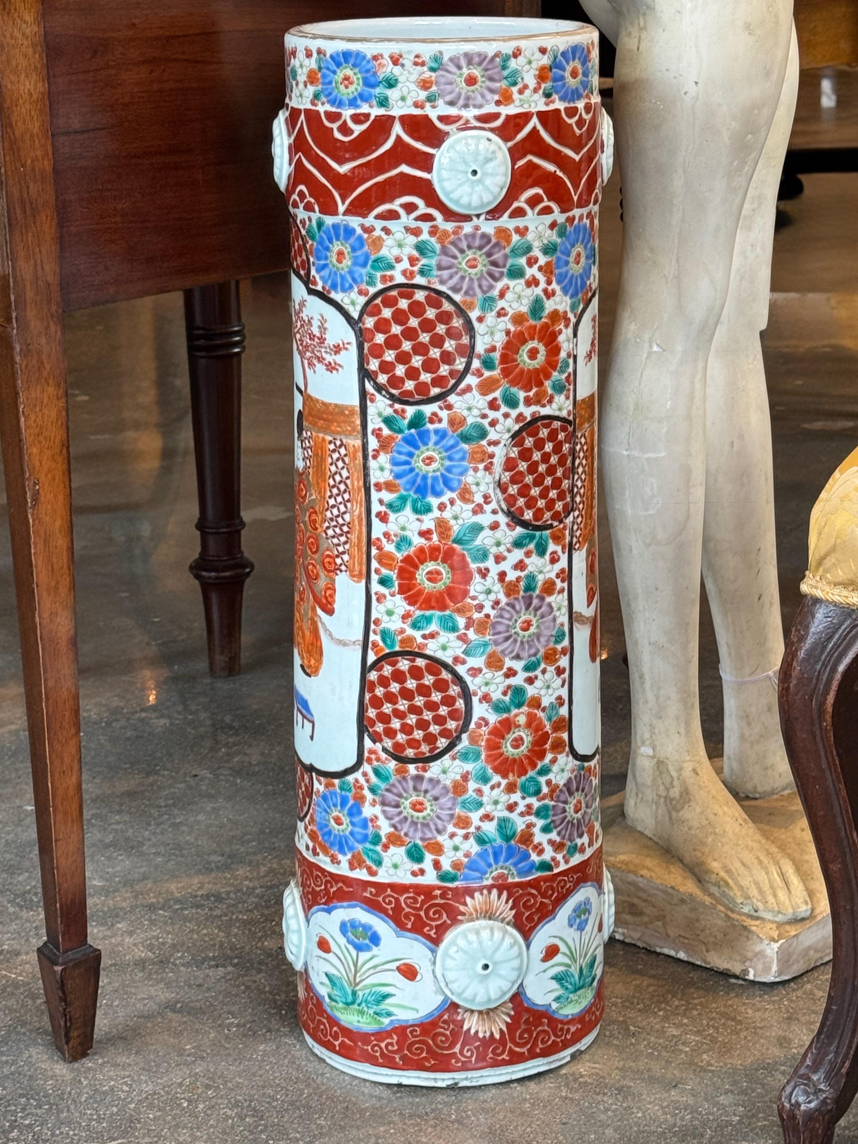 Grand porte-parapluie cylindrique en porcelaine avec une décoration vibrante de style Imari dans des tons rouges, bleus, verts et lavande. La surface est ornée de panneaux de motifs floraux et géométriques, bordés de médaillons en relief. Une pièce