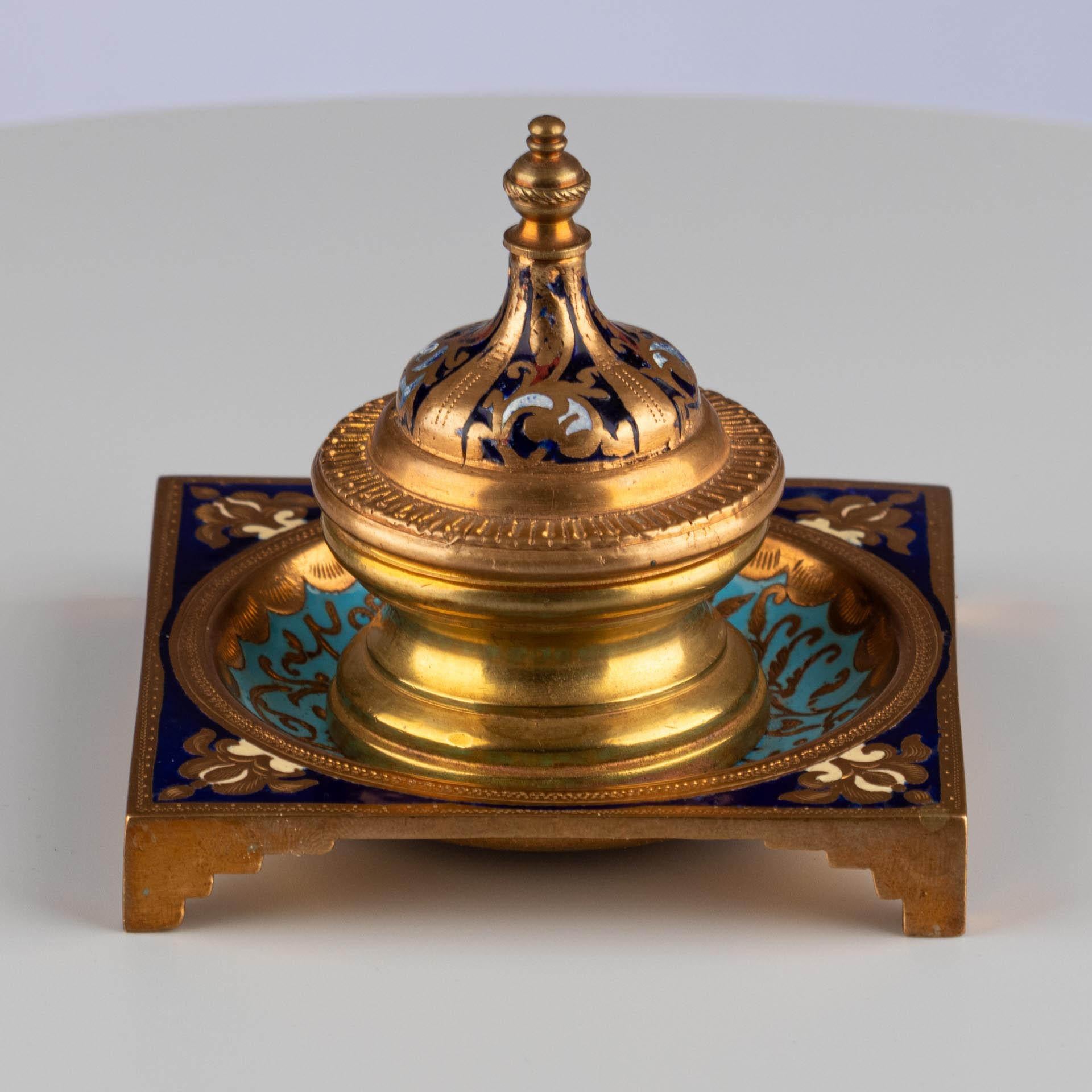 Calamaio con decorazione a smalto

Origine: Francia
Materiale: Metallo dorato con smalto colorato
Data: Circa 1870-1890

Dimensioni:
H. H. × L. × P.: 8 × 9 × 9 cm.

Descrizione:
Calamaio quadrato appoggiato su quattro piedi a gradini, con un