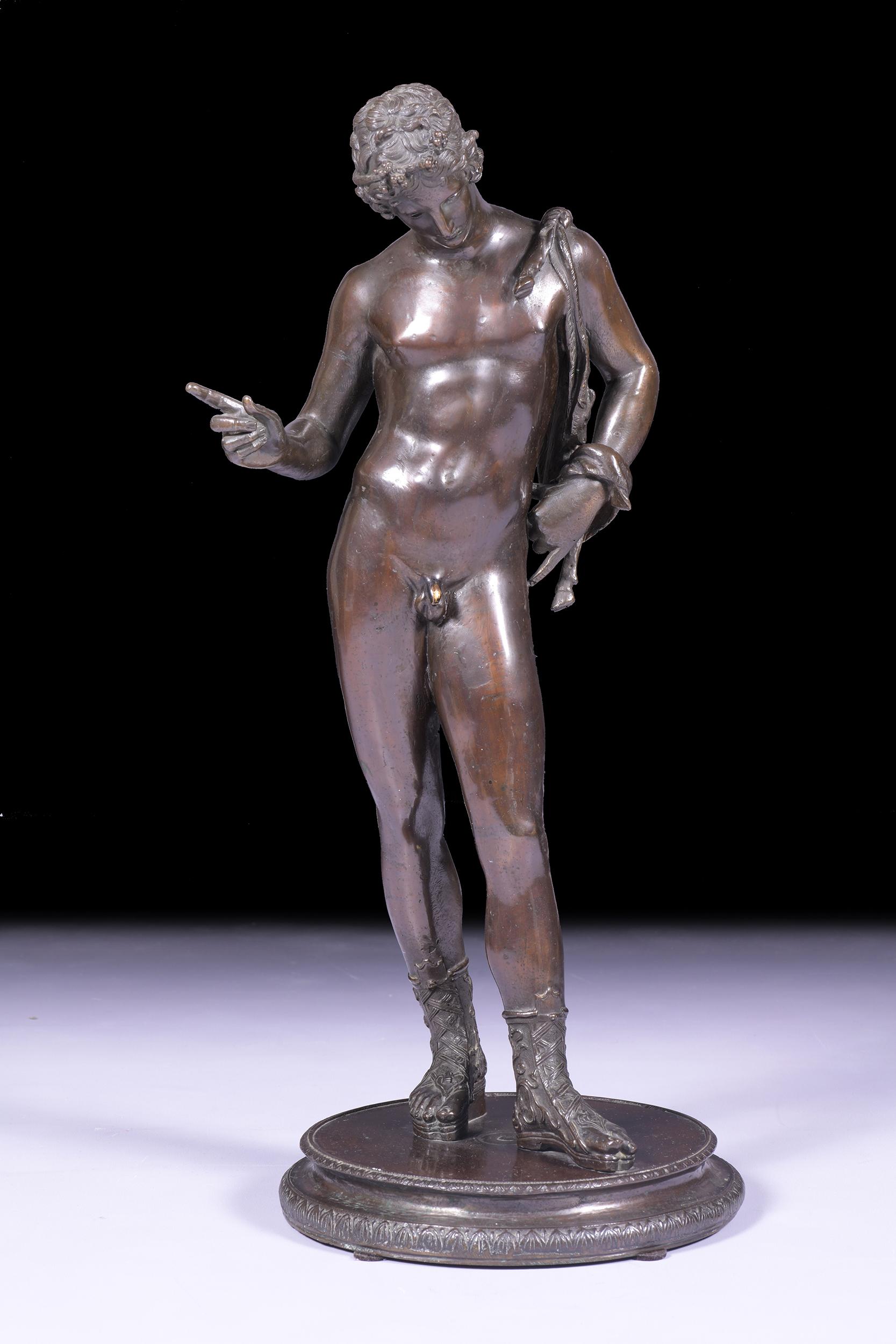 A 19th century bronze figure of Narcissus, after the Antique, the standing nude wearing a wreath, one sandal clad foot before the other, his right hand pointing upwards, his left shoulder slung with a goats skin wine sack, on circular base with