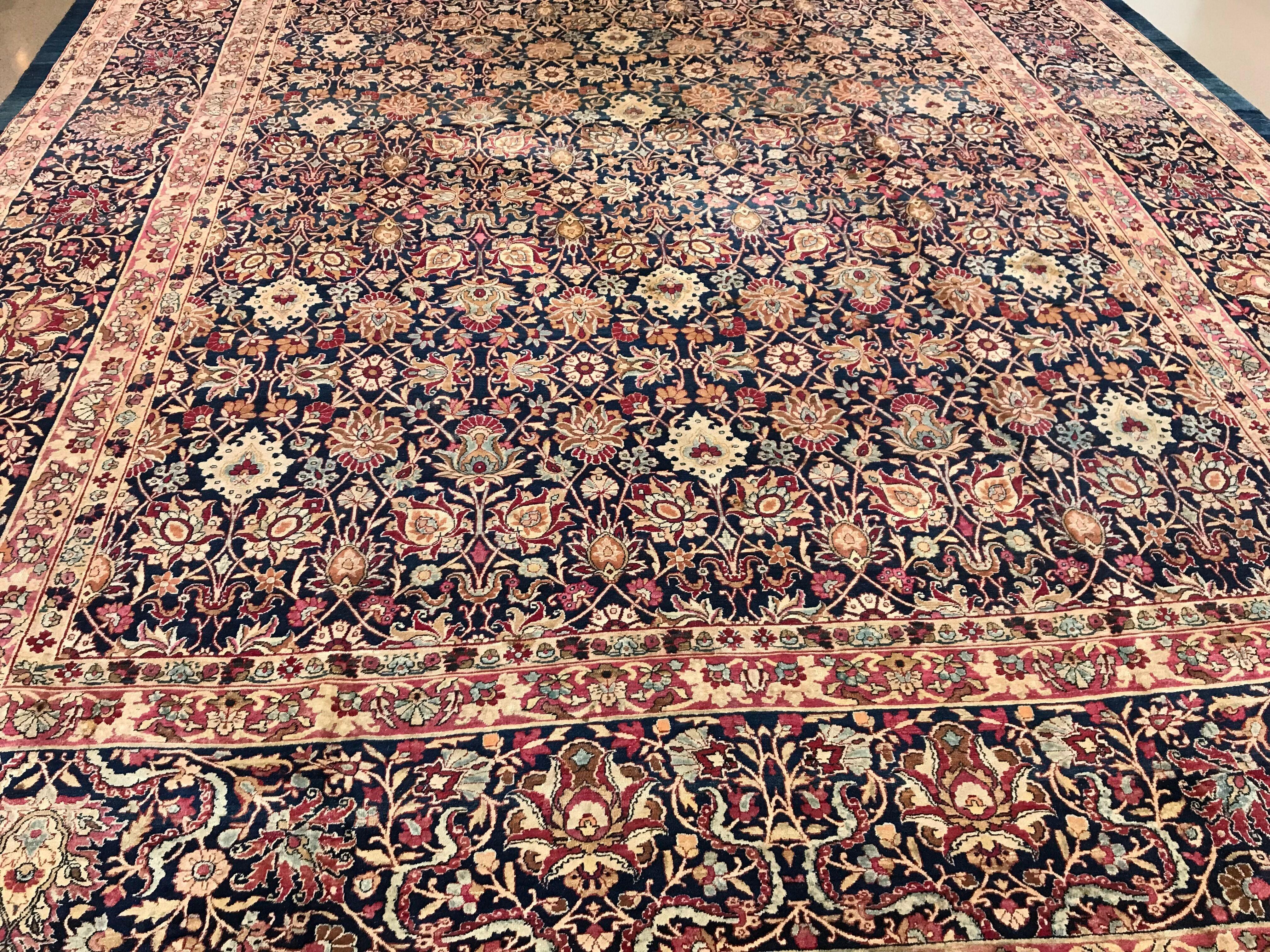 Ce tapis persan traditionnel tissé à la main à Kerman présente un champ indigo profond composé de palmettes ornementées et de vignes angulaires formant un treillis en losange, dans une majestueuse bordure indigo composée de palmettes audacieuses et