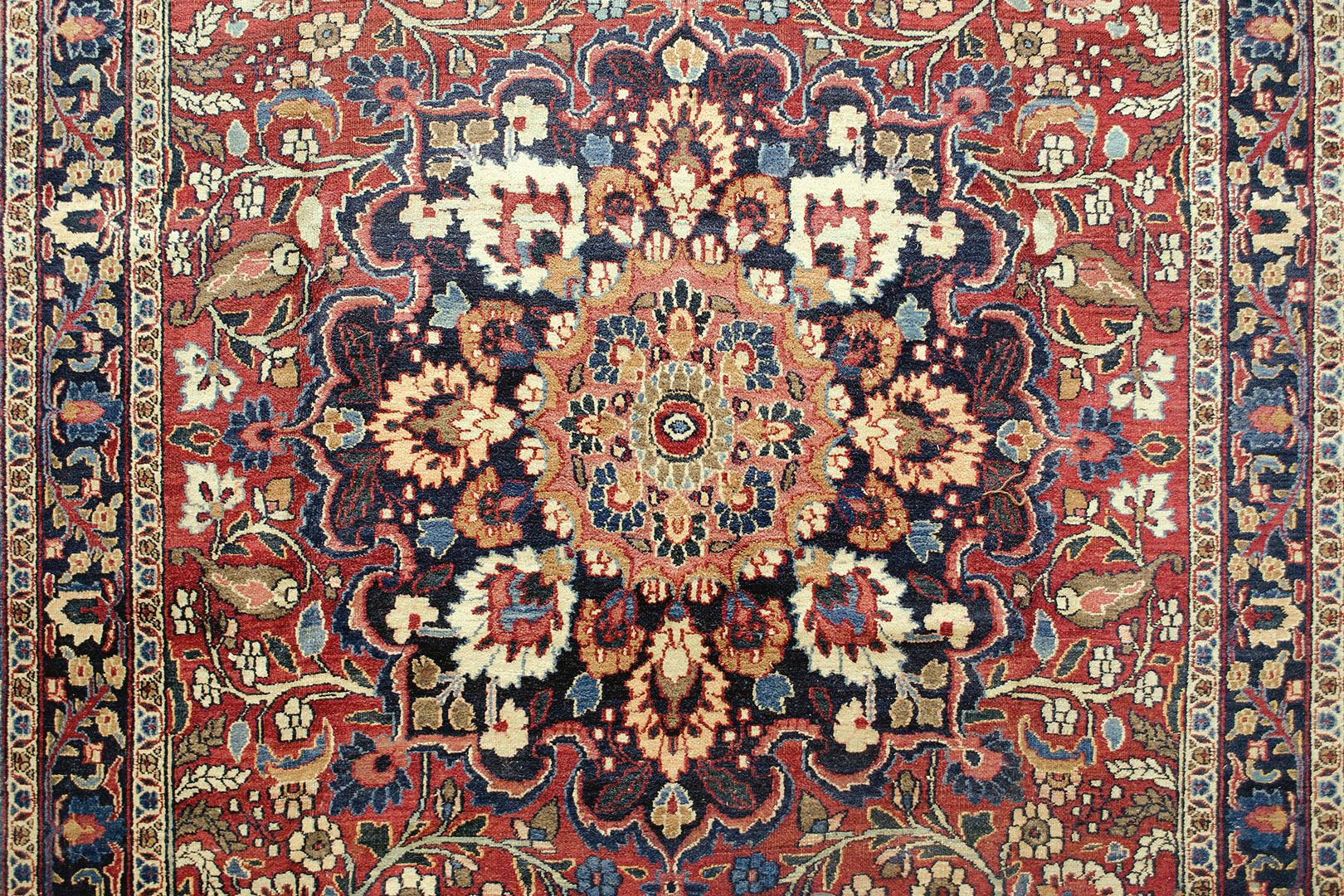 Ce tapis persan traditionnel tissé à la main présente un champ rubis vibrant de vignes florales à enroulement dense entourant un majestueux médaillon arabesque polychrome, avec des écoinçons de palmettes saphir, dans une bordure de palmettes saphir