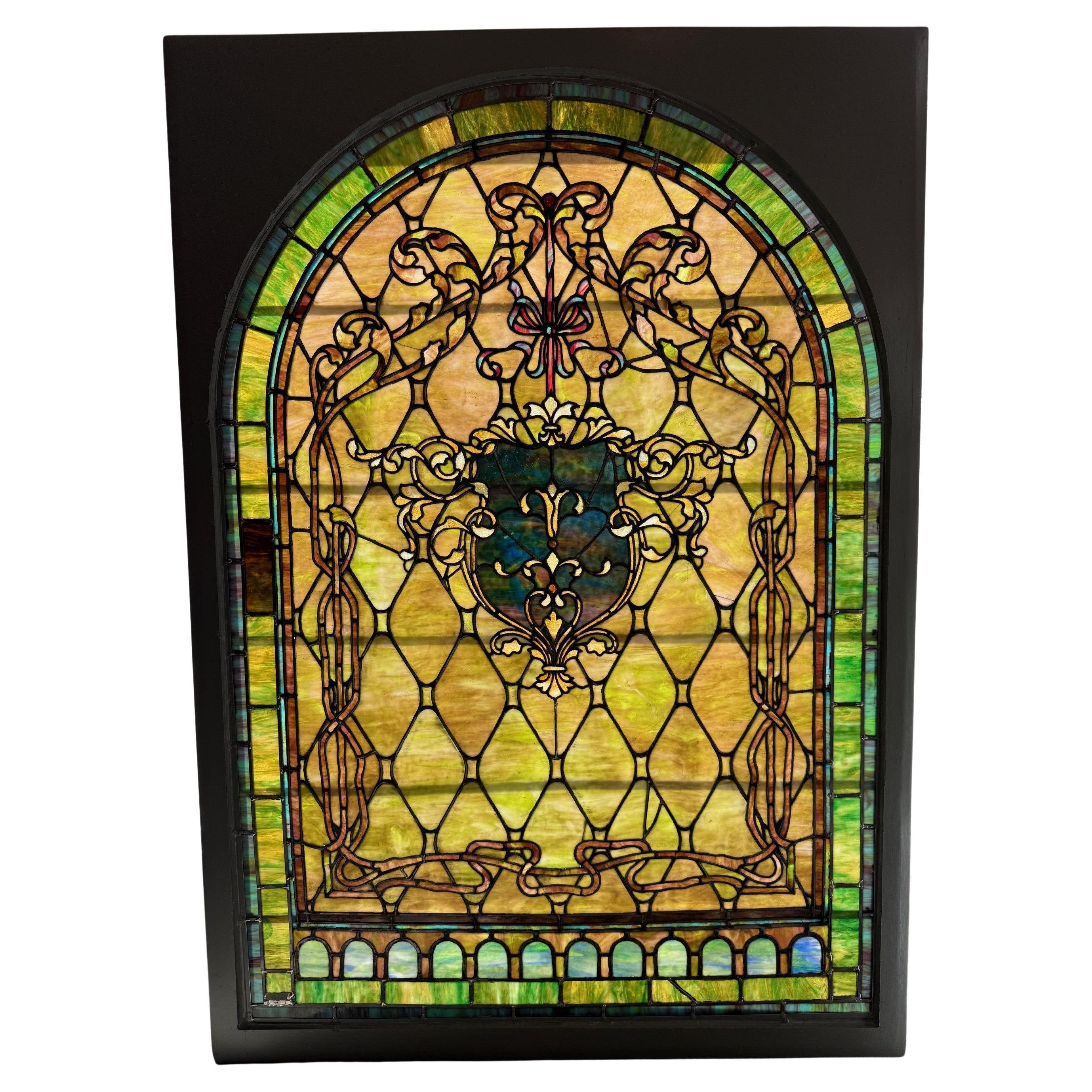 Gran vidriera arqueada de finales del siglo XIX con marco de madera. Se trata de una hermosa ventana con grandes colores, cintas decorativas y un escudo en el centro. Recuperada de una gran finca y reparada con paneles de colores a juego, es un