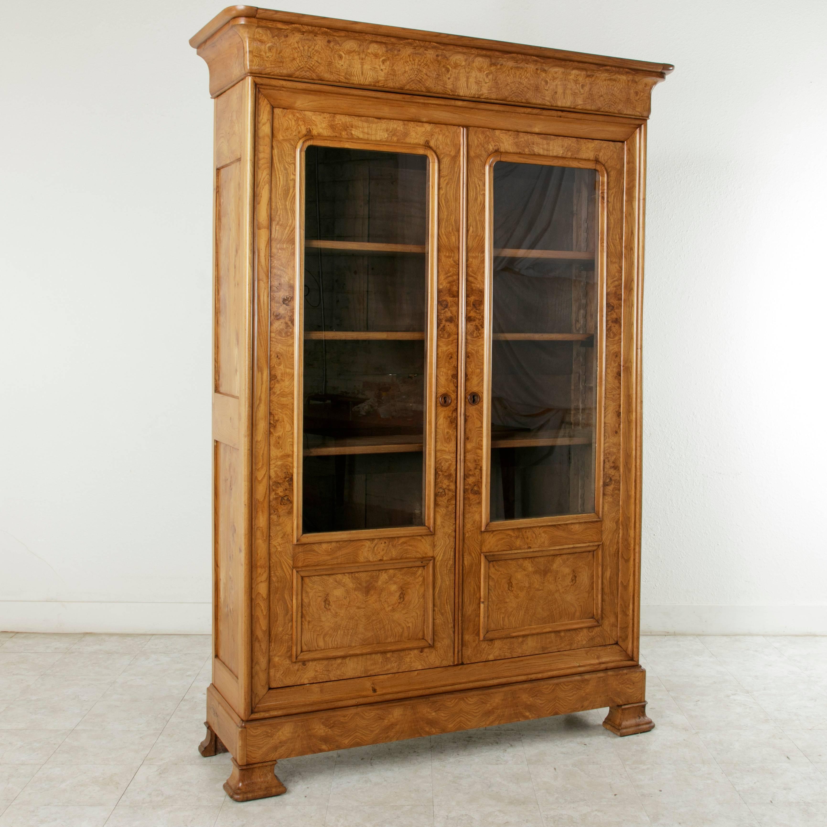 This Louis Philippe style bibliothèque or bookcase from the late 19th century features a facade of exquisite book matched burl ash. Two doors inset with glass open to reveal an interior with four adjustable shelves that provide ample storage and