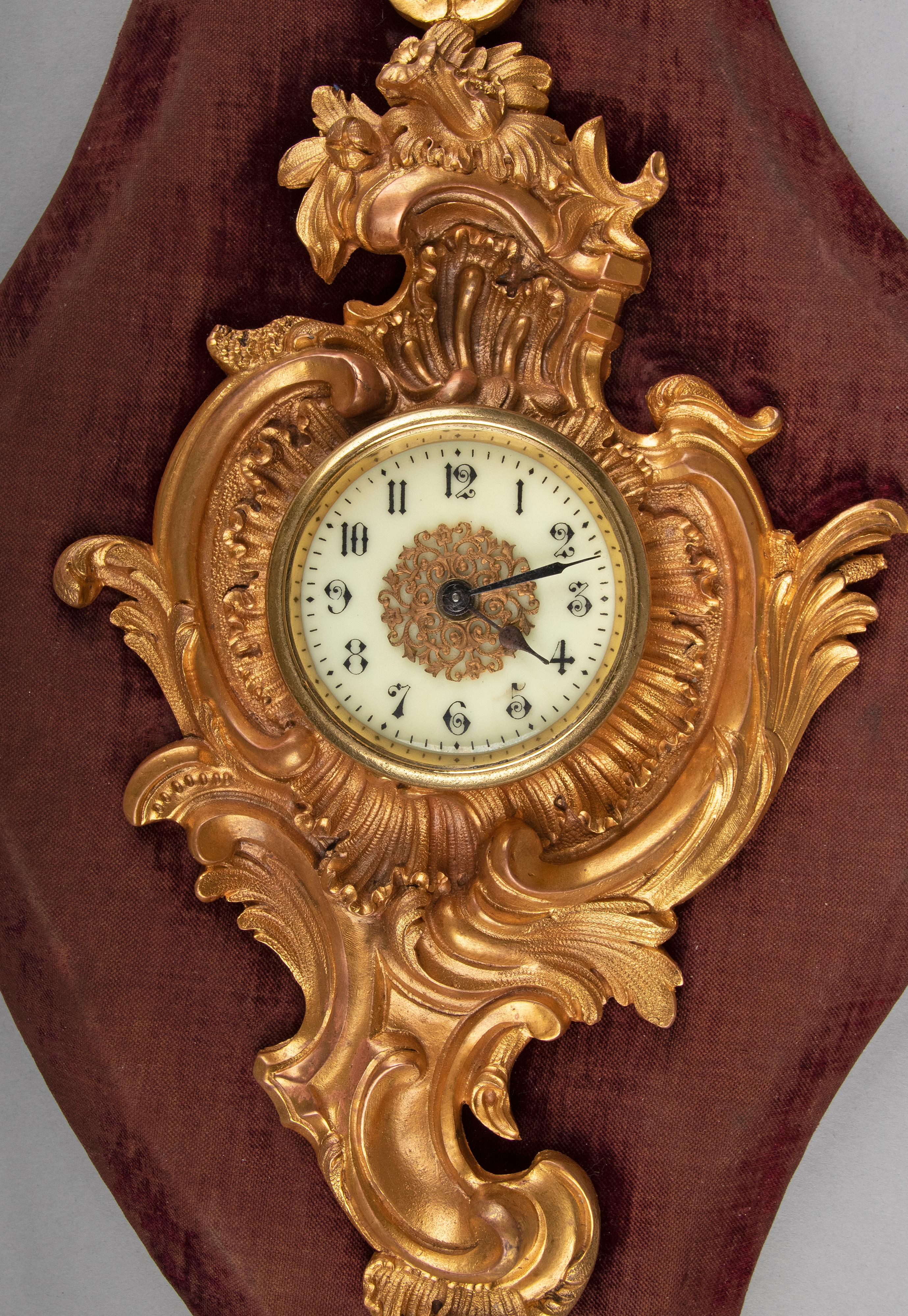 A small and elegant antique cartel clock, mounted on a wooden base with fabric. The richly decorated clock is made of bronze with refined details. Ornated with Louis XV style Rococo scrolls. The dial is made of enameled copper with Arabic numbers.