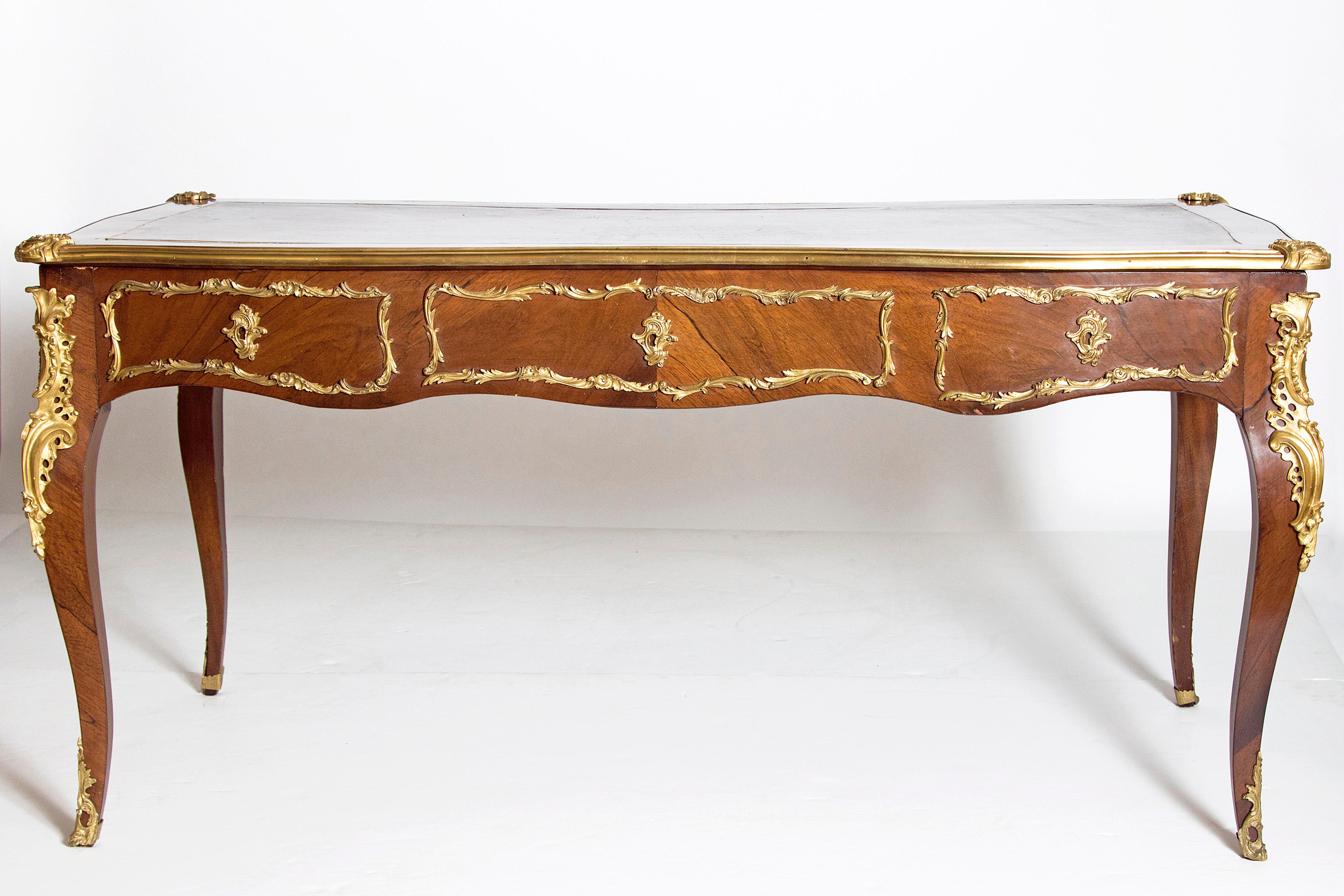 A elegant French late 19th century Louis XV style rosewood and ormolu bureau plat. The top is a gilt tooled shaped leather fitted within a veneer border surrounded by an ormolu galley with foliate ormolu mounts at each corner. Below the frieze