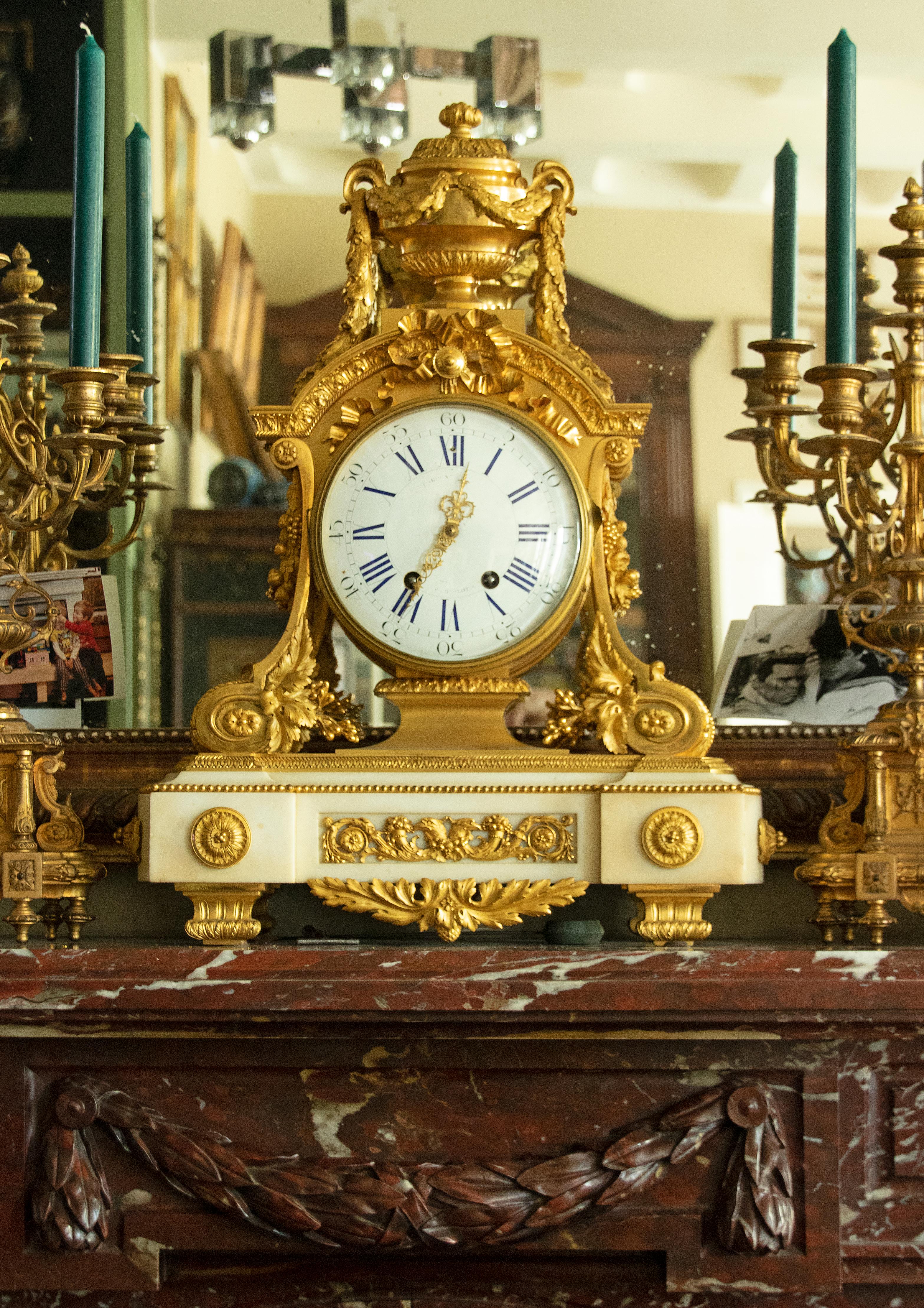 A large and richly decorated ormolu bronze mantel clock on a white carrara marble base. The clock is embellished all around with bronze ornaments in Louis XVI style; a lidded coupe with a garland of oak leaf on top, tied bow and floral motives. The