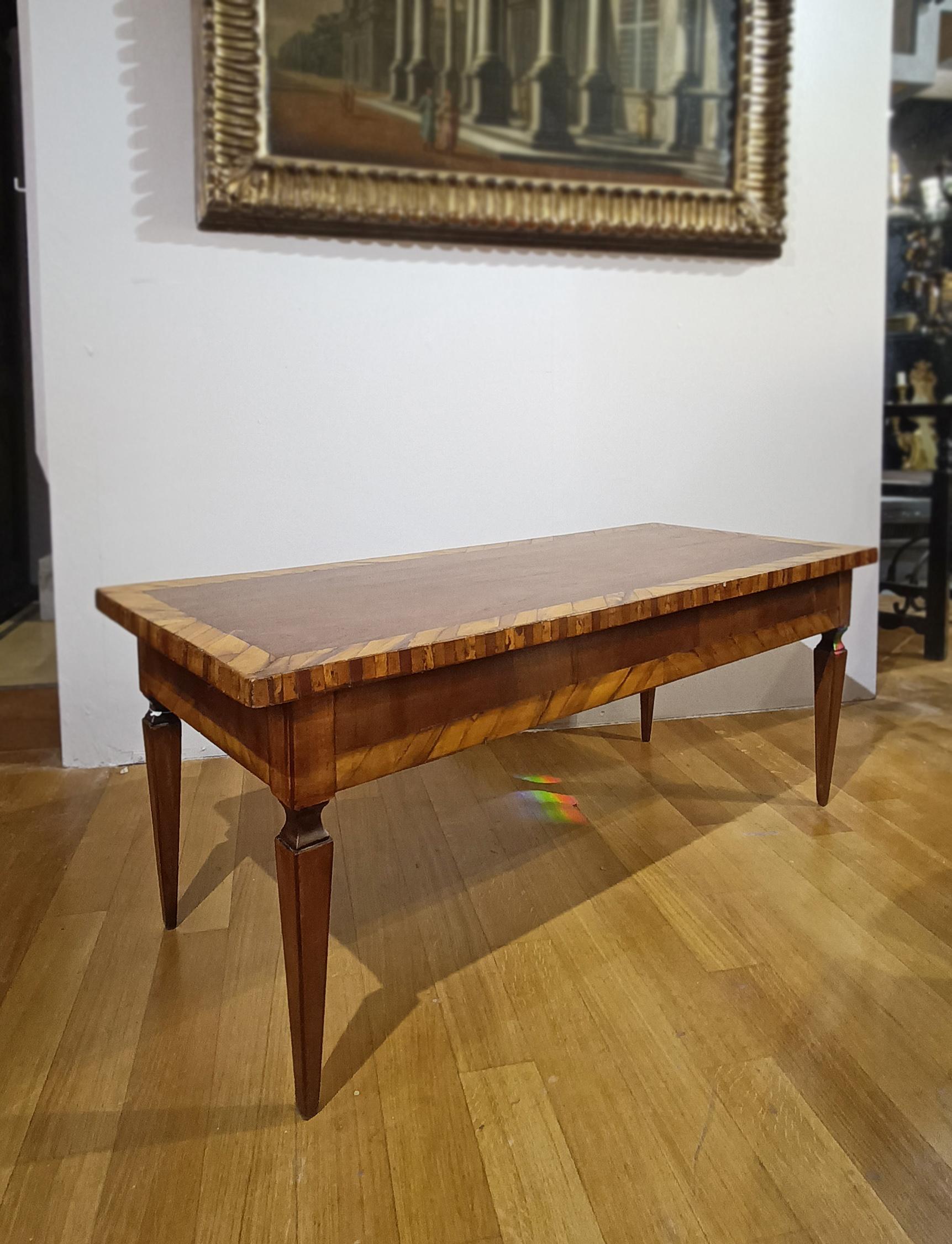 Elégante table basse de style Louis XVI, fabriquée en noyer massif sculpté et agrémentée de placages de noyer et d'olivier. La structure se distingue par ses quatre pieds pyramidaux tronqués et les placages détaillés sur le dessus et les côtés, qui